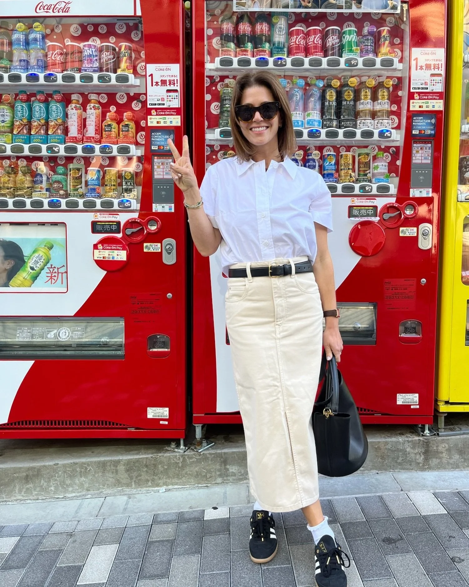 Person posing in front of drink vending machines, wearing sunglasses, white shirt, beige skirt, and holding a black bag.