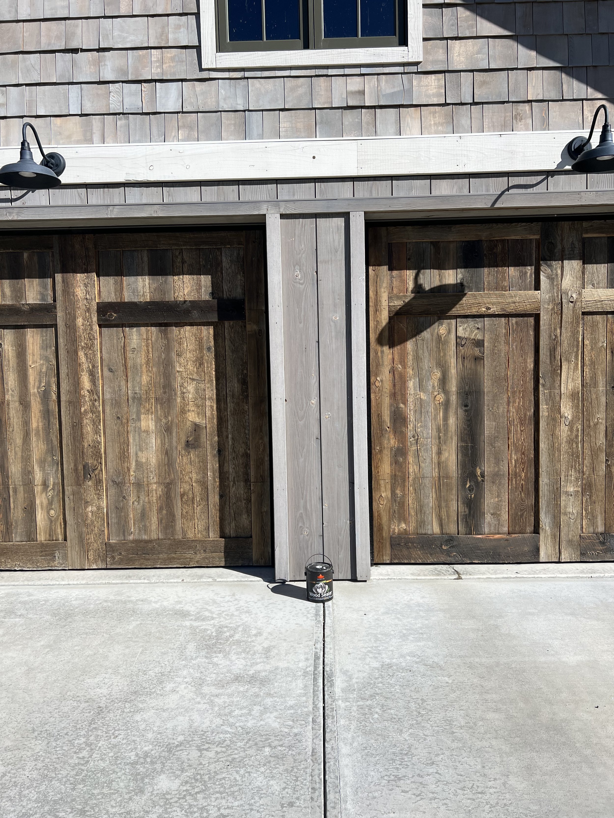 Two wooden garage doors with shingle siding and outdoor light fixtures; a small decorative lantern is on the ground in the sunlight.