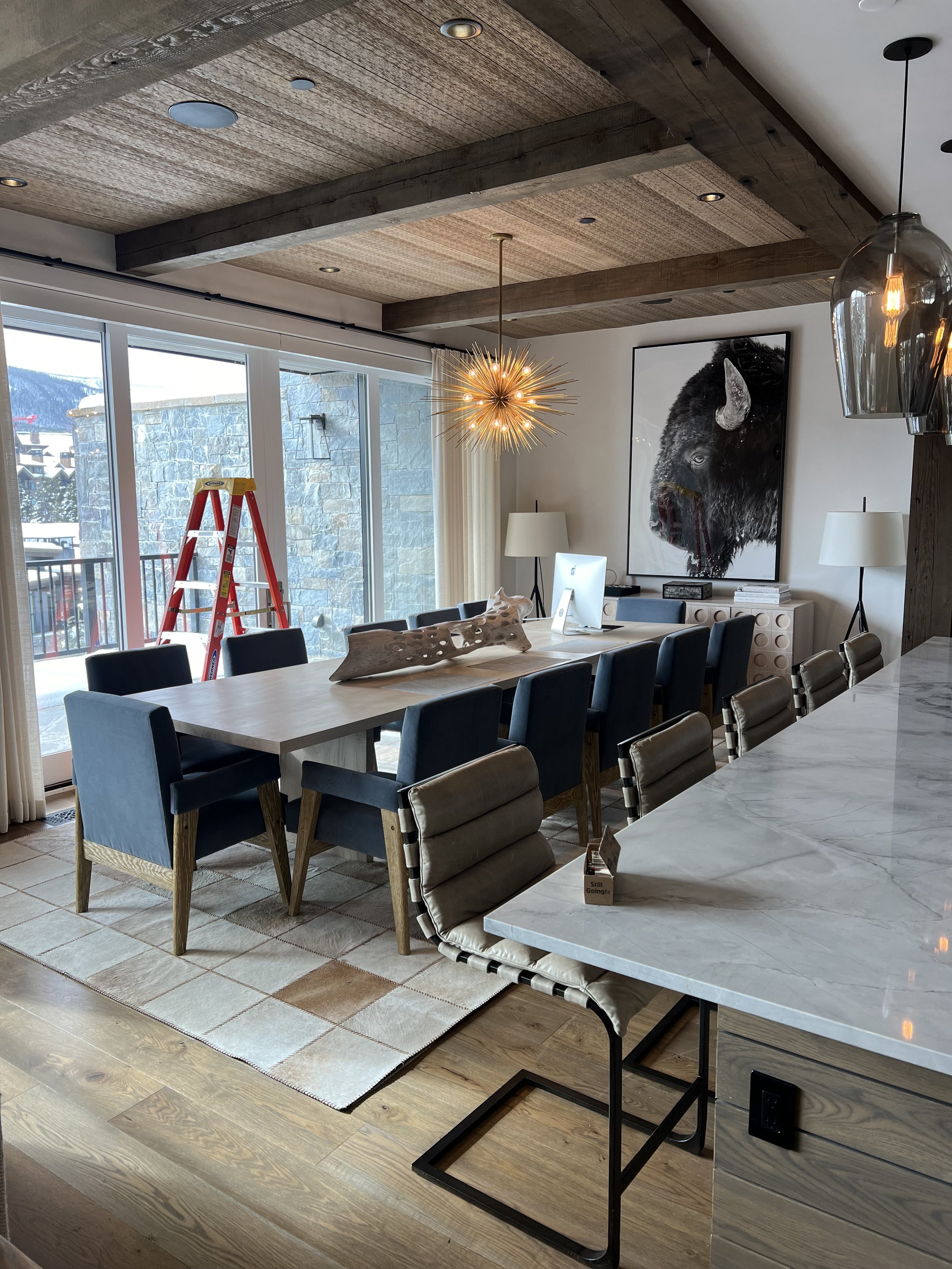 Modern dining room with a long table, blue chairs, and a unique light fixture. A large window, wall art of a bison, and a step ladder are visible.