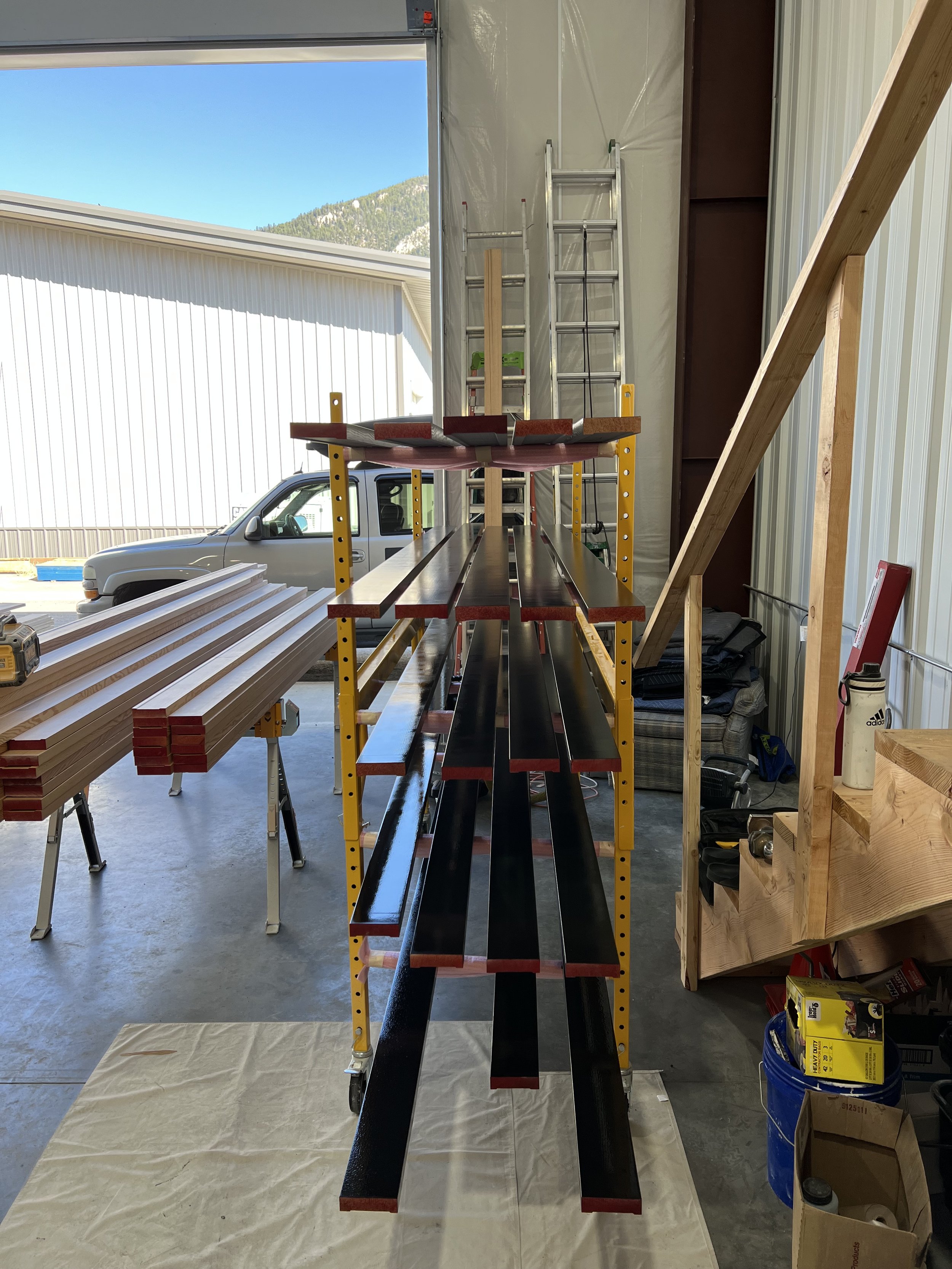 Interior view of a workshop with wooden planks on a yellow shelving unit, ladders against the wall, and an open garage door showing an outdoor structure and a vehicle.