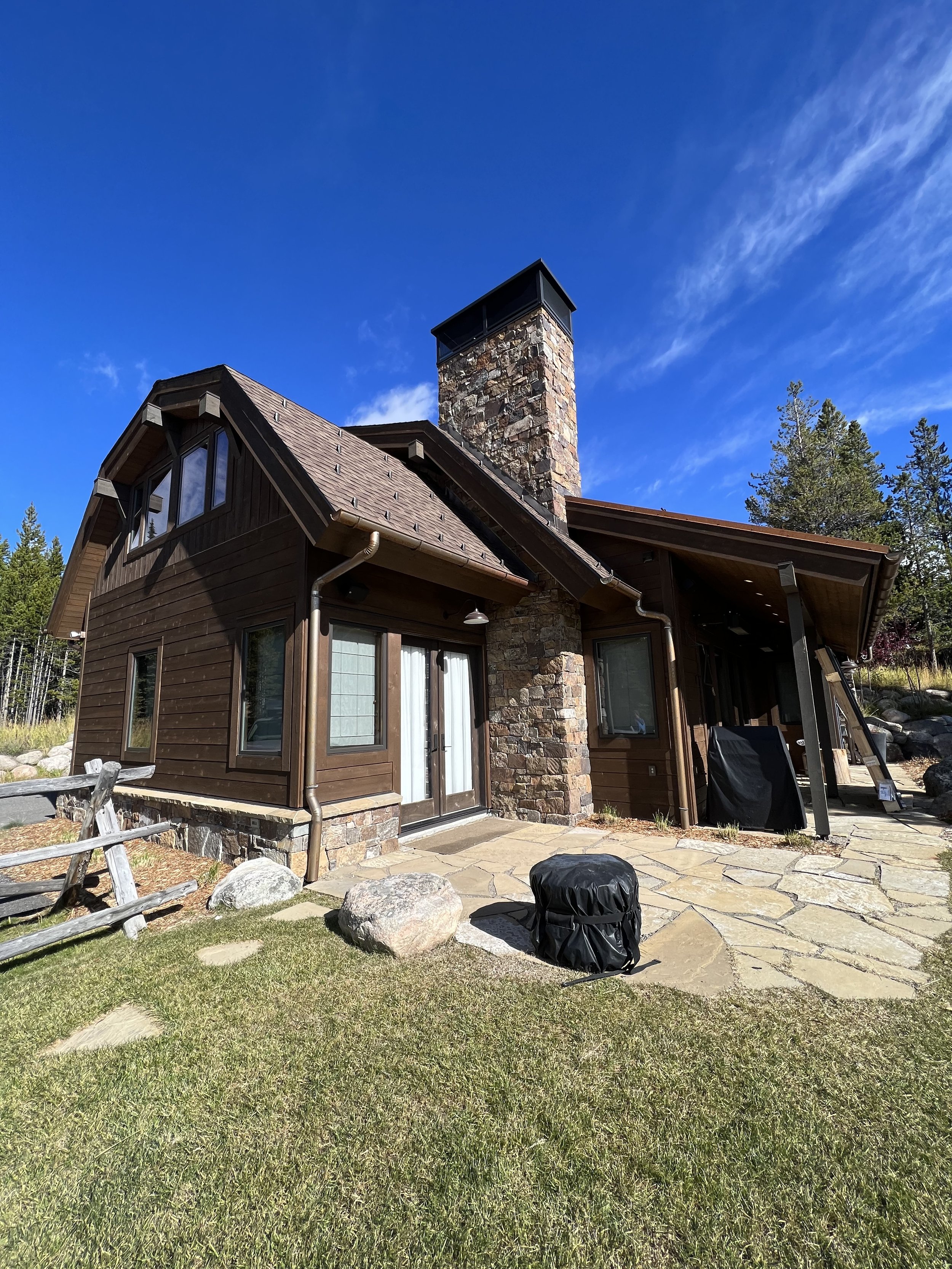 Rustic wooden house with stone chimney and patio in a grassy outdoor setting.
