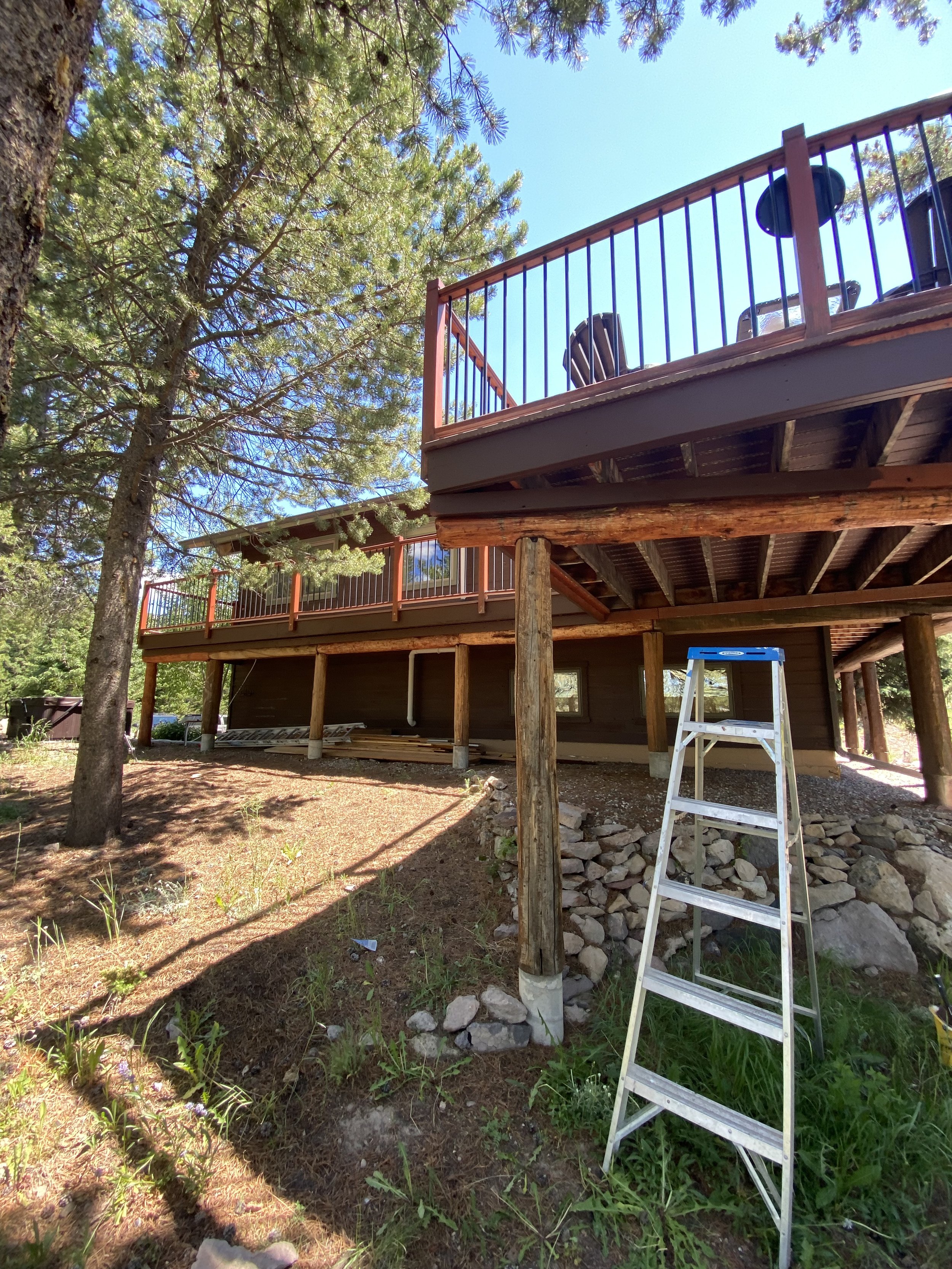 Two-story wooden house with a raised deck, surrounded by trees. A ladder leans against the house.