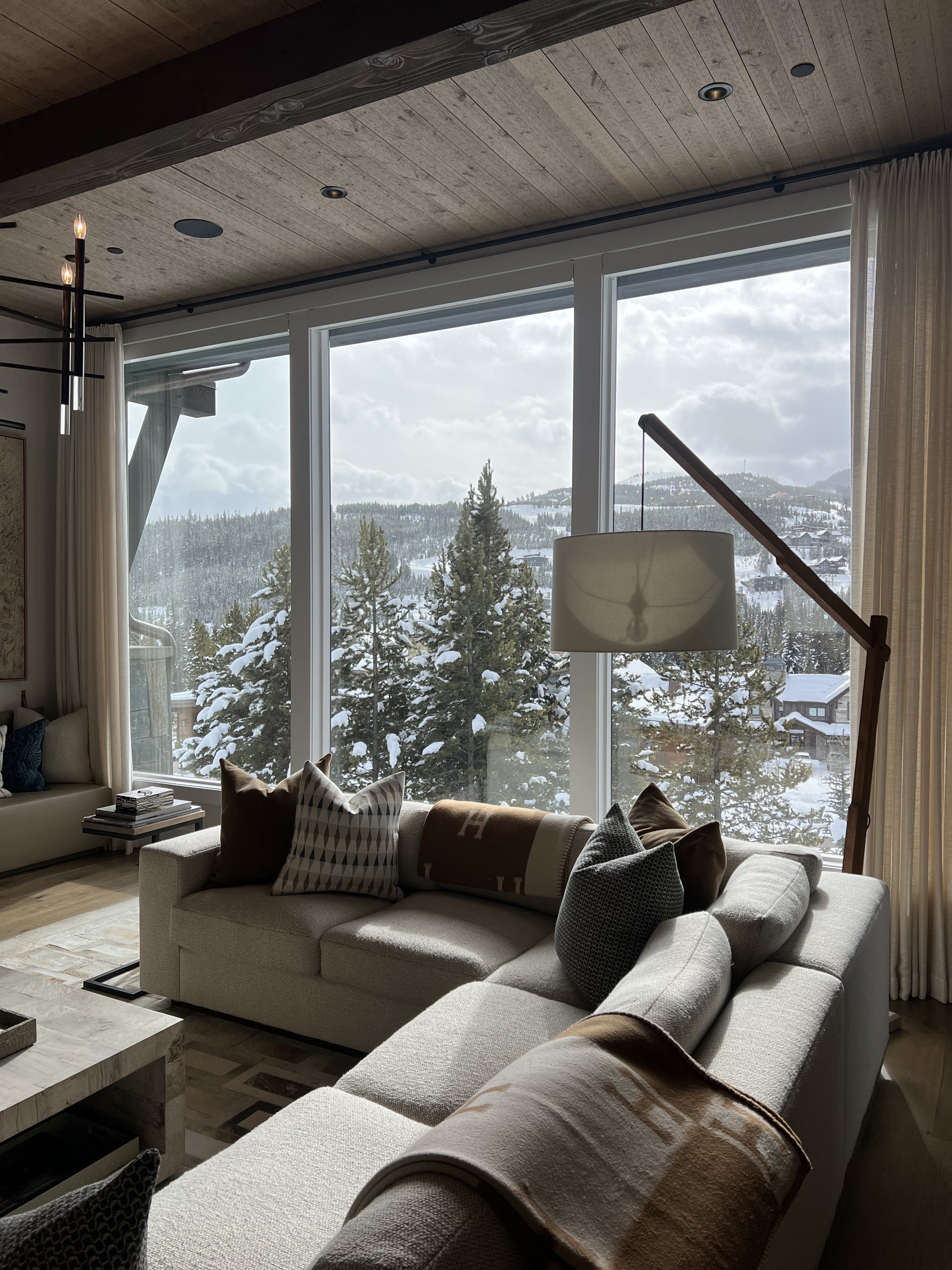 Cozy living room with large windows showcasing a snow-covered outdoor view, featuring a sectional sofa with pillows and blankets, a wooden coffee table, and stylish floor lamp.