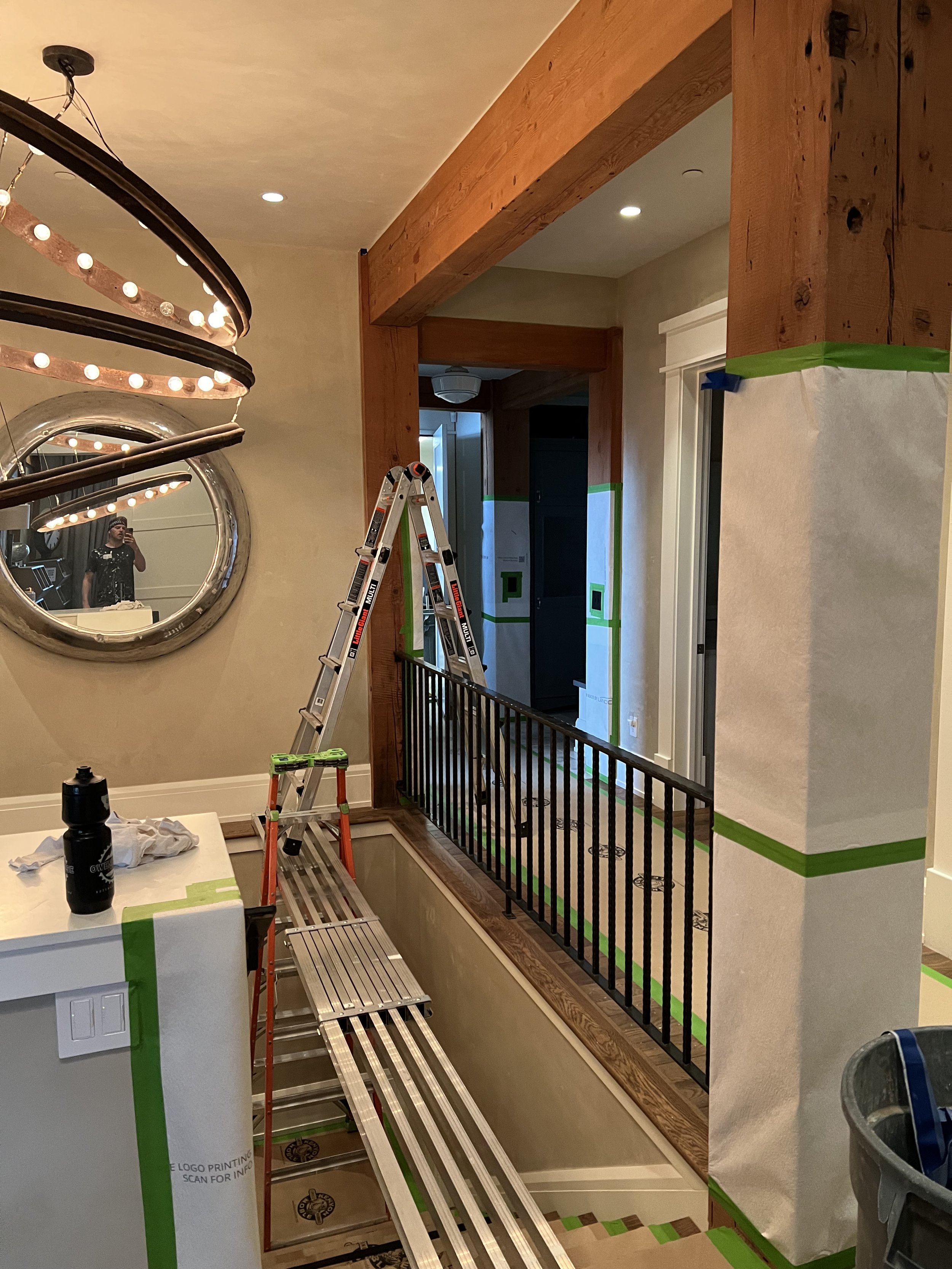 Indoor staircase under renovation, featuring a metal scaffold ladder bridge across the stairs, large wooden beams, a circular mirror, and a modern chandelier with exposed bulbs. Walls and surfaces are covered with protective material for painting or