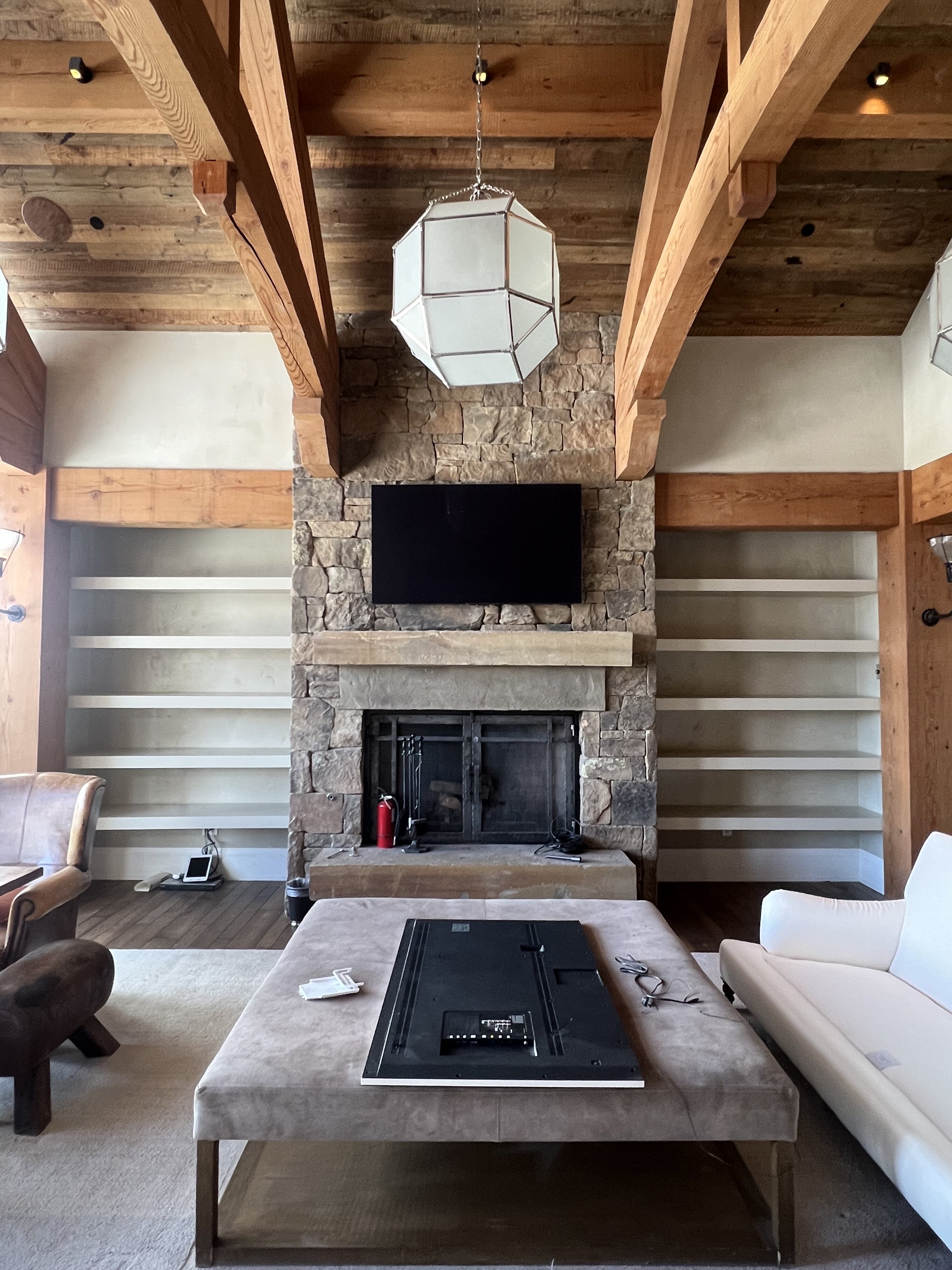 Rustic living room with stone fireplace, large wooden beams, and a modern geometric light fixture. It features empty shelves, a flat-screen TV, a white sofa, and a brown coffee table with cables and a device on it.