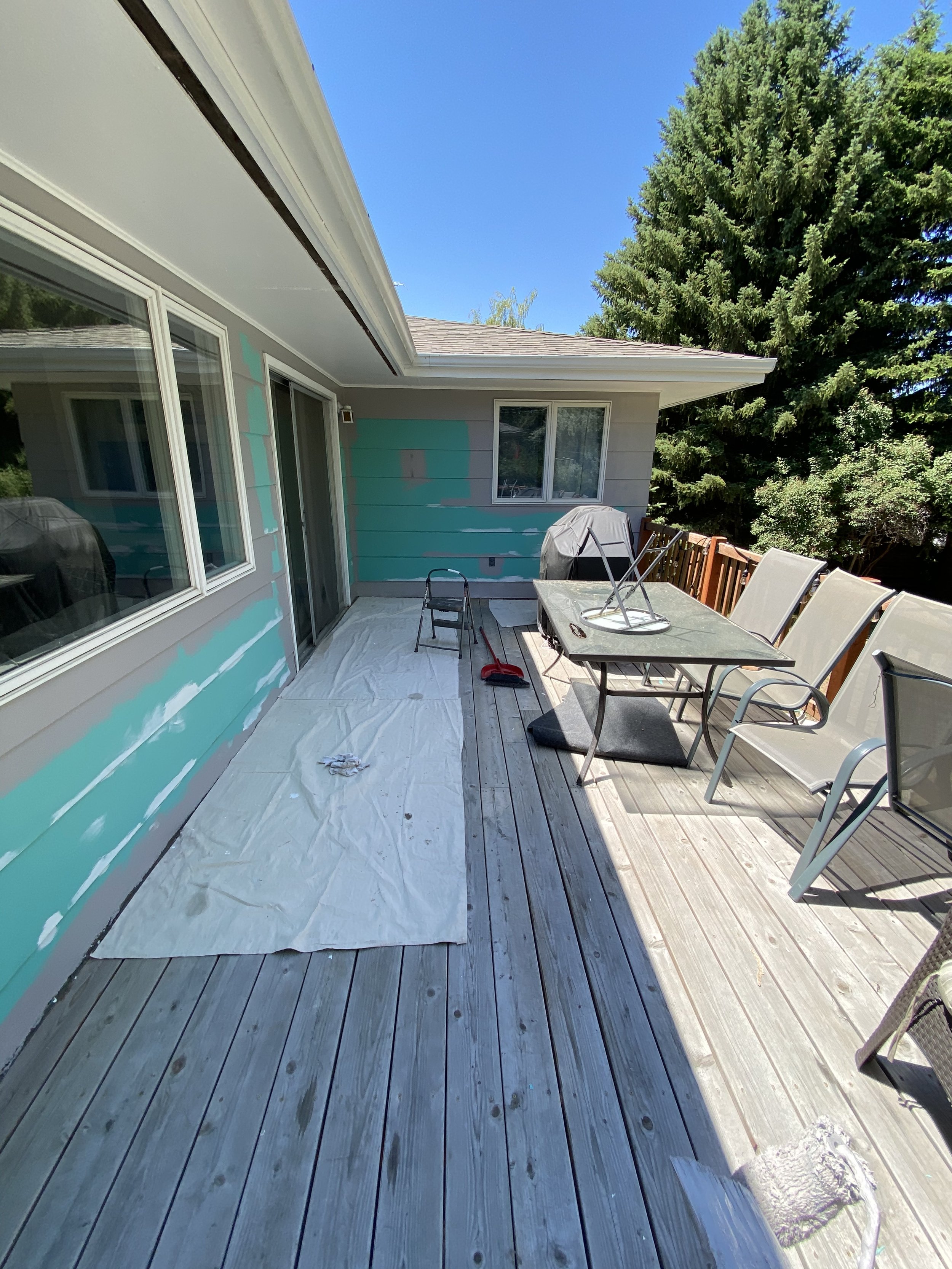 Wooden deck with a table, chairs, grill, and painting supplies on drop cloths, adjacent to a house with partially painted siding, surrounded by trees.