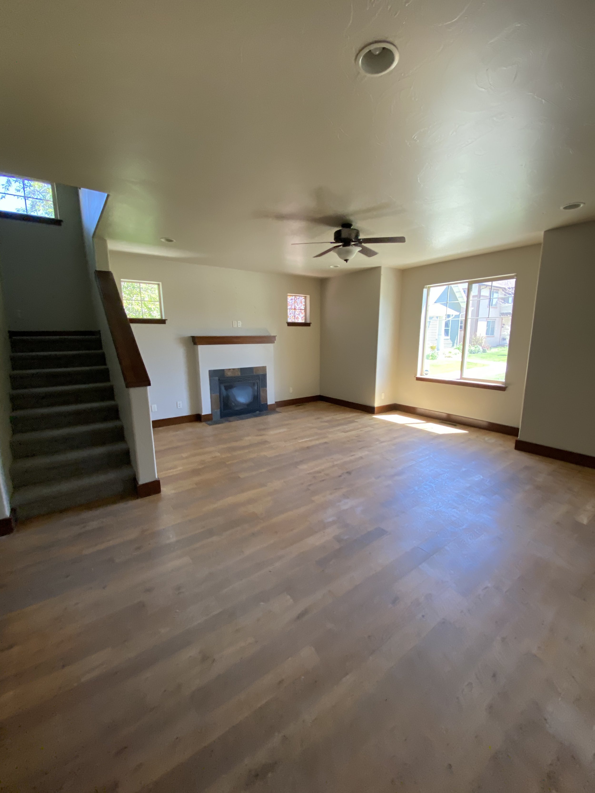 Empty living room with wooden floors, staircase, fireplace, ceiling fan, and large window.