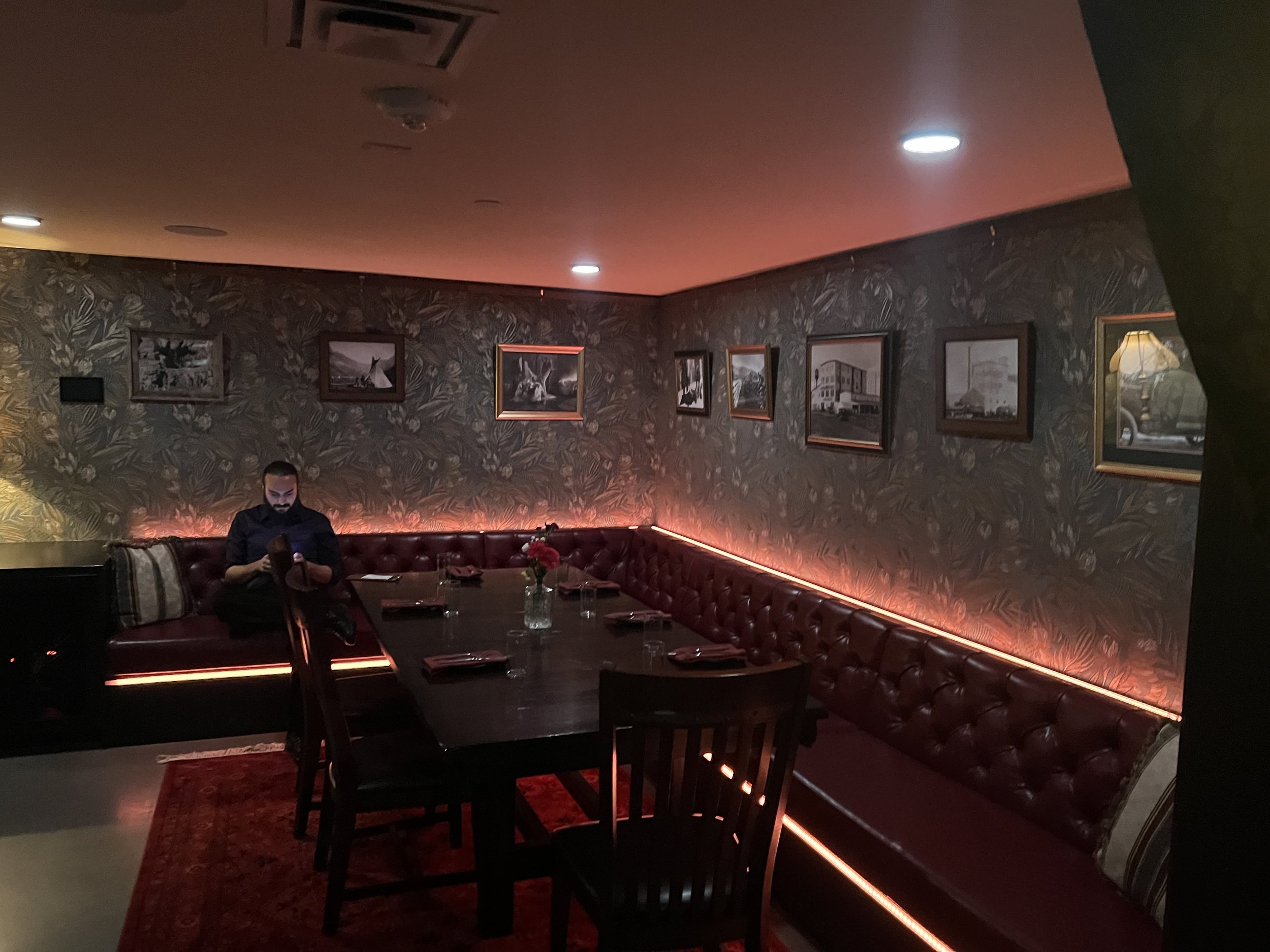 Dimly lit restaurant interior with leather booths, framed pictures on floral wallpaper, and a man using a smartphone at a dark wooden table.