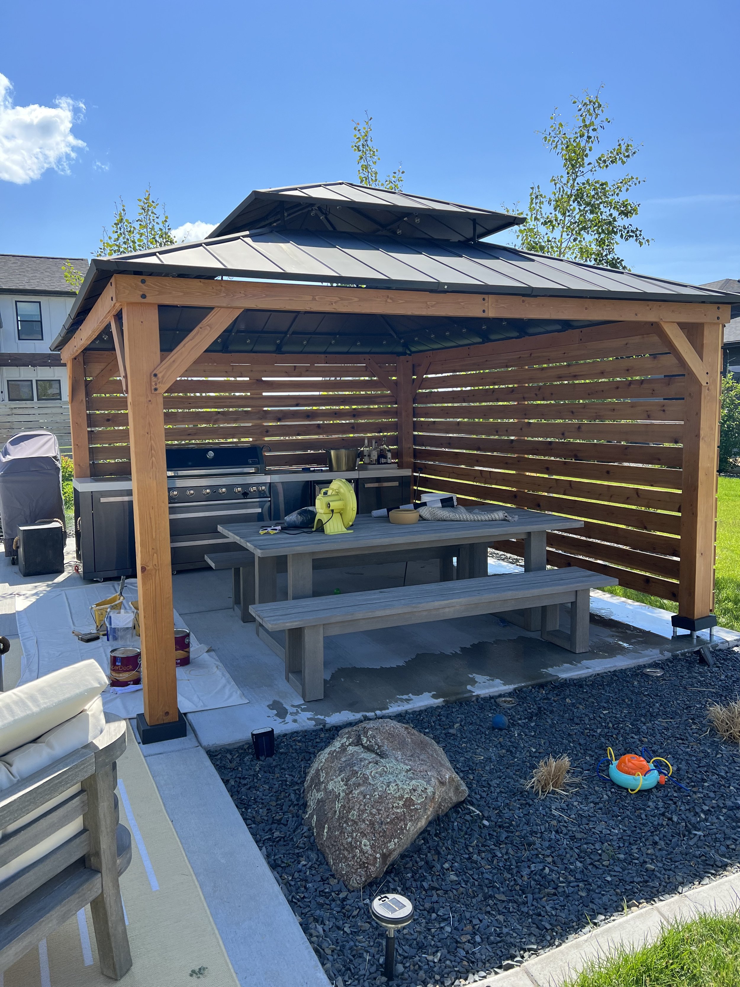 Outdoor gazebo with wooden structure and metal roof, housing a grill and picnic table. The area has a landscaped rock garden and children's toy on gravel. Sunny day with clear blue sky.