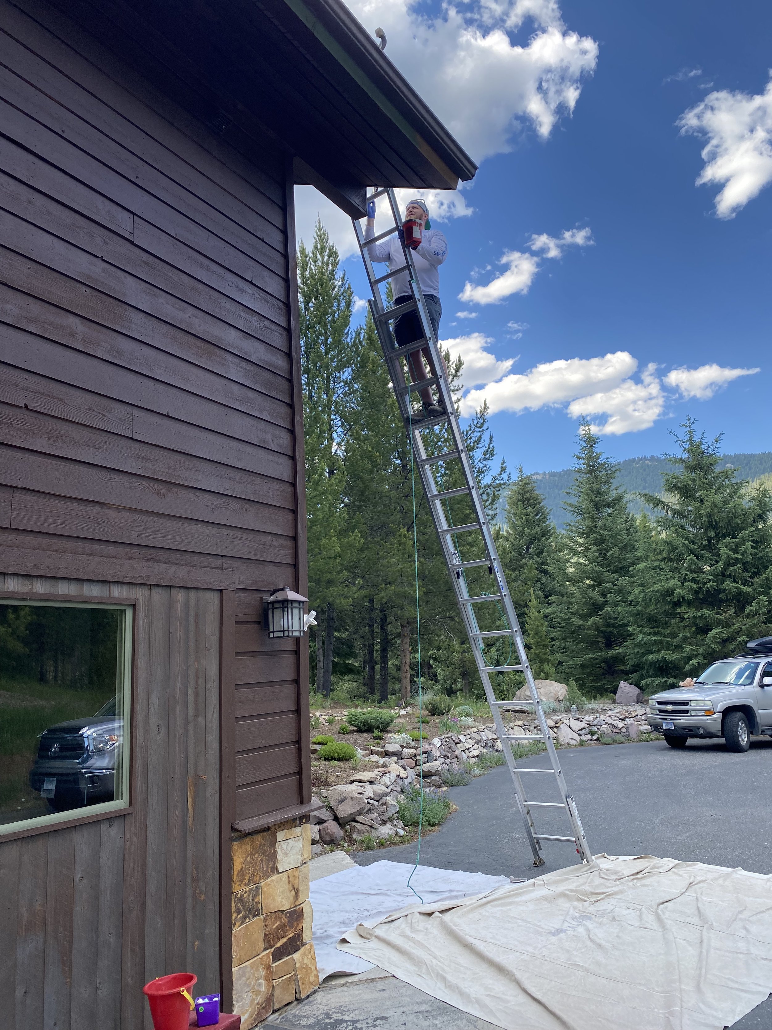 Person on ladder painting house, surrounded by trees, vehicles, and drop cloth, under a blue sky with clouds.