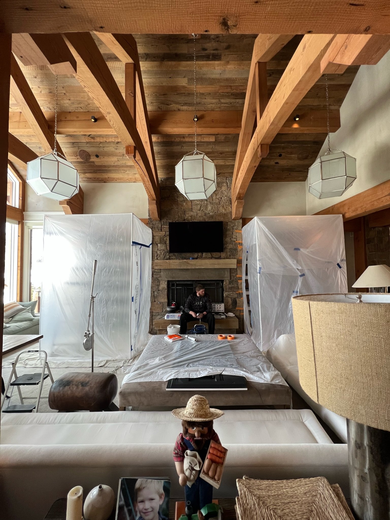 Living room interior with exposed wooden beams, covered furniture for renovation, and a person sitting near a fireplace. Hanging geometric lights and a rustic decor style are visible.