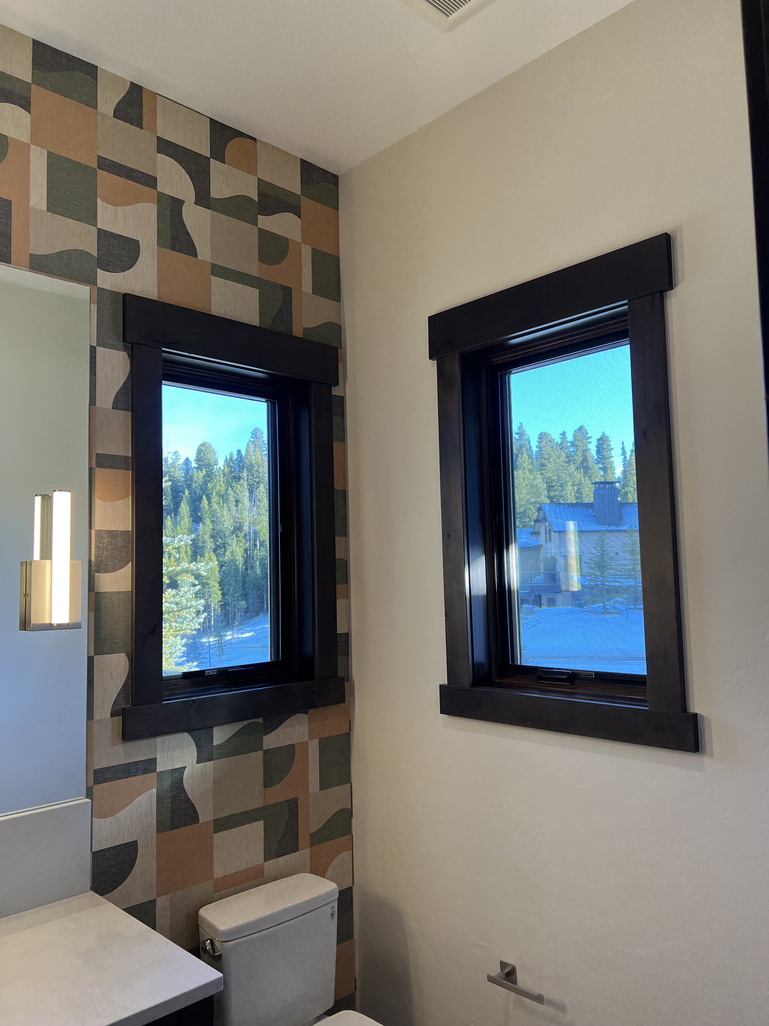 Bathroom interior with abstract patterned wallpaper, a white toilet, two windows with dark frames, a rectangular mirror, and a modern wall sconce. The view outside shows trees and a building.