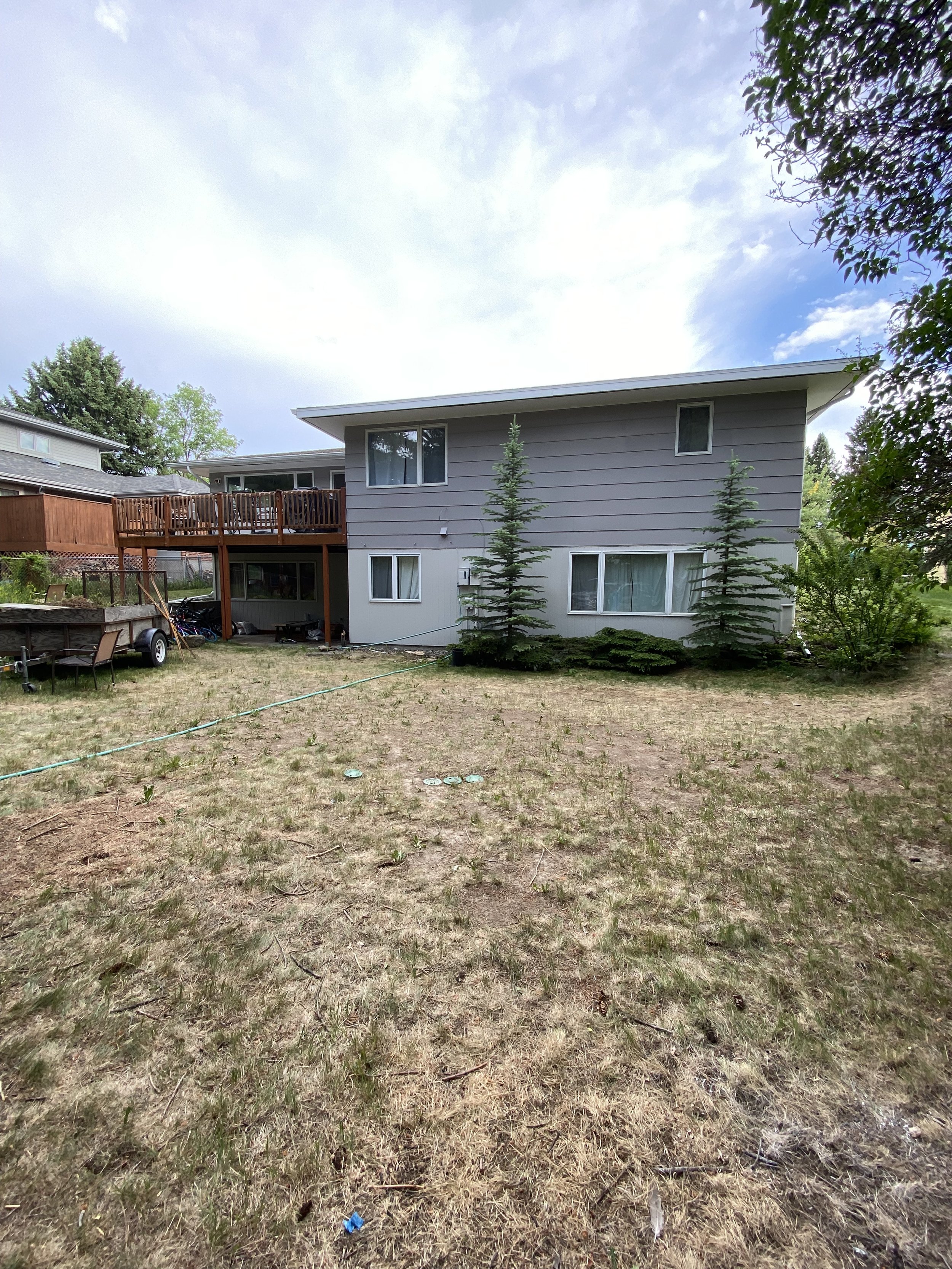 Two-story house with gray siding, elevated wooden deck, sparse grassy yard, and several tall trees nearby.