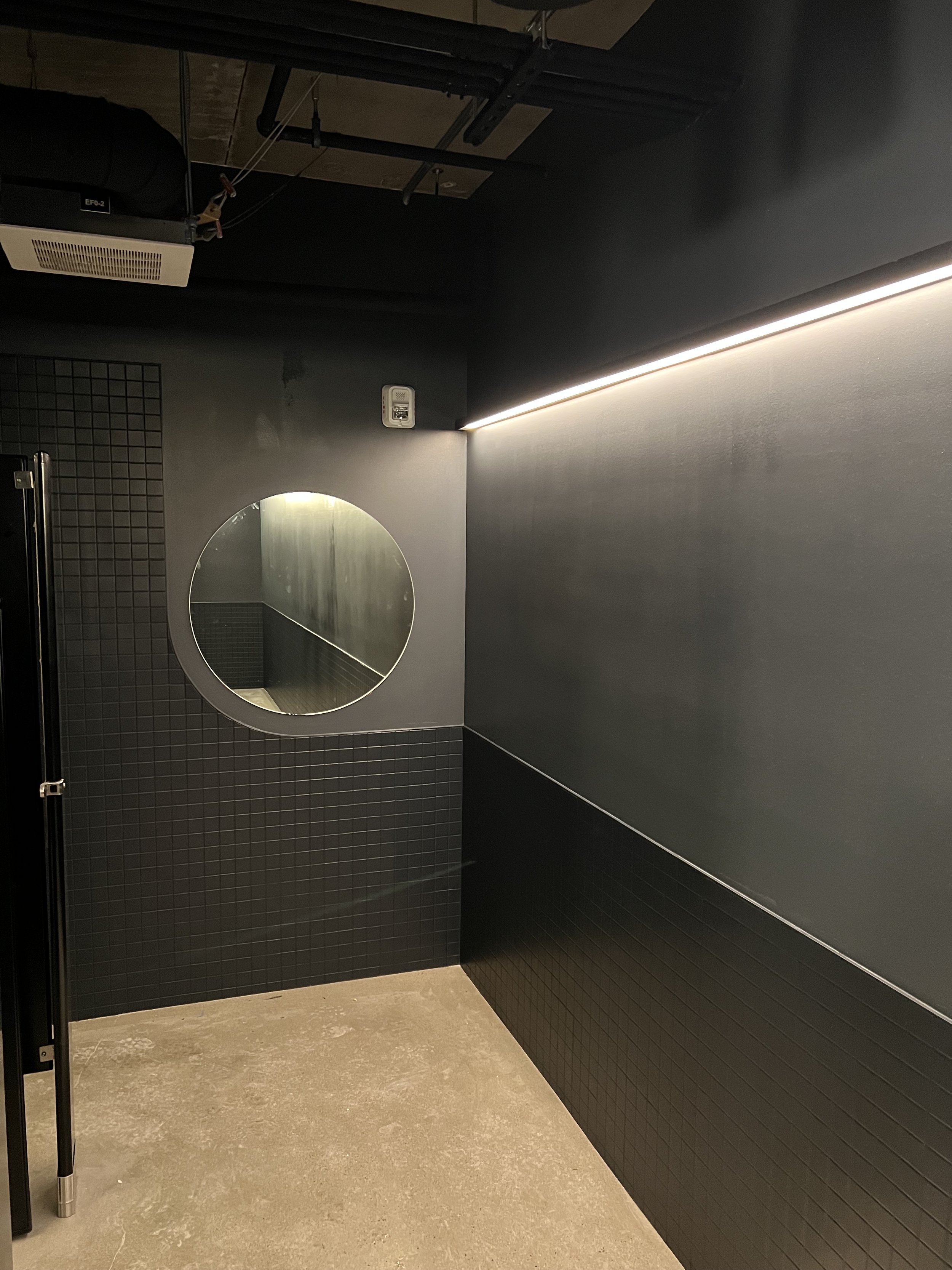 Modern restroom interior with dark tiles, round mirror, concrete floor, and overhead lighting.