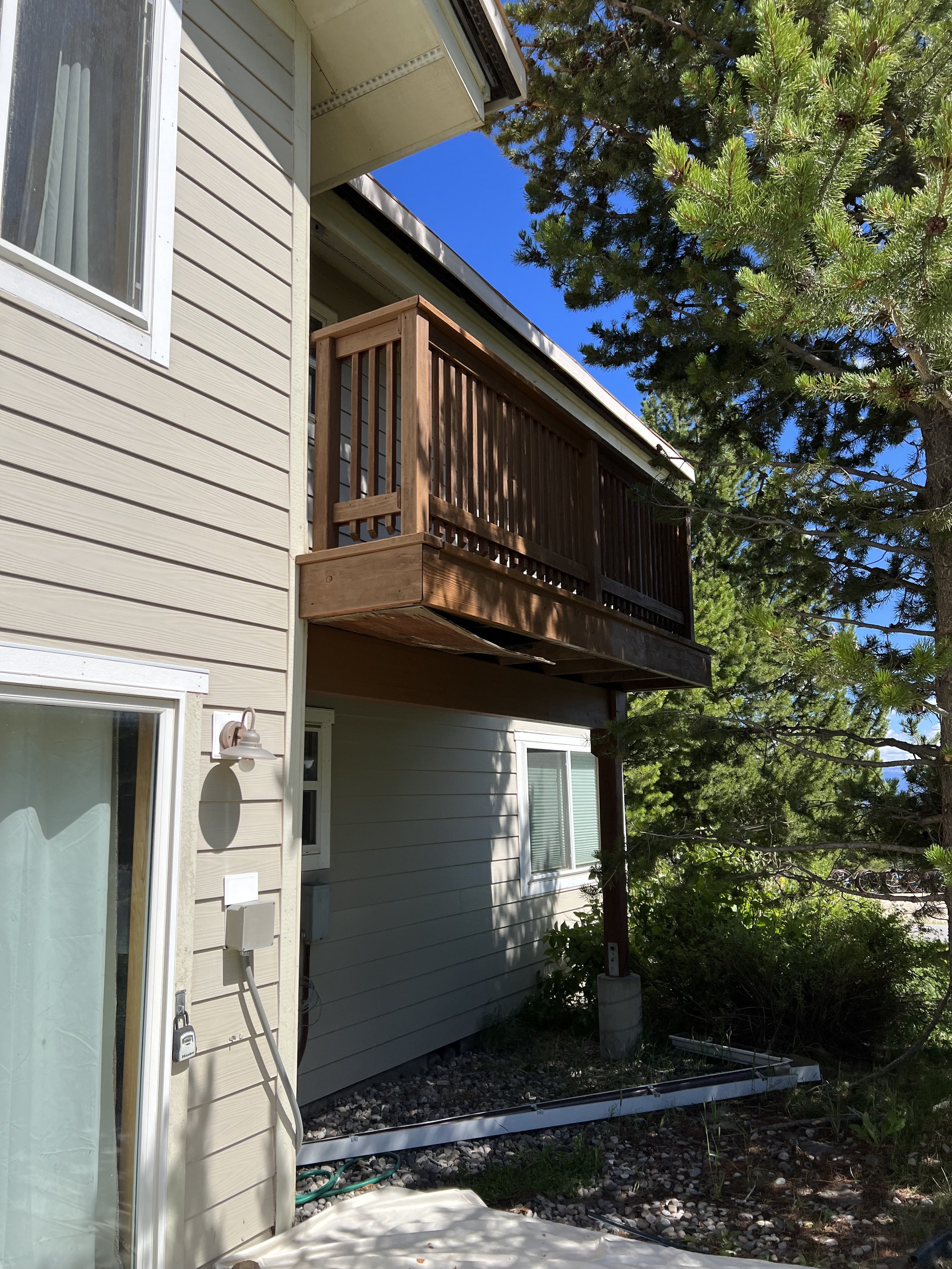 Exterior view of a beige house with a small wooden balcony, surrounded by pine trees and shrubs.