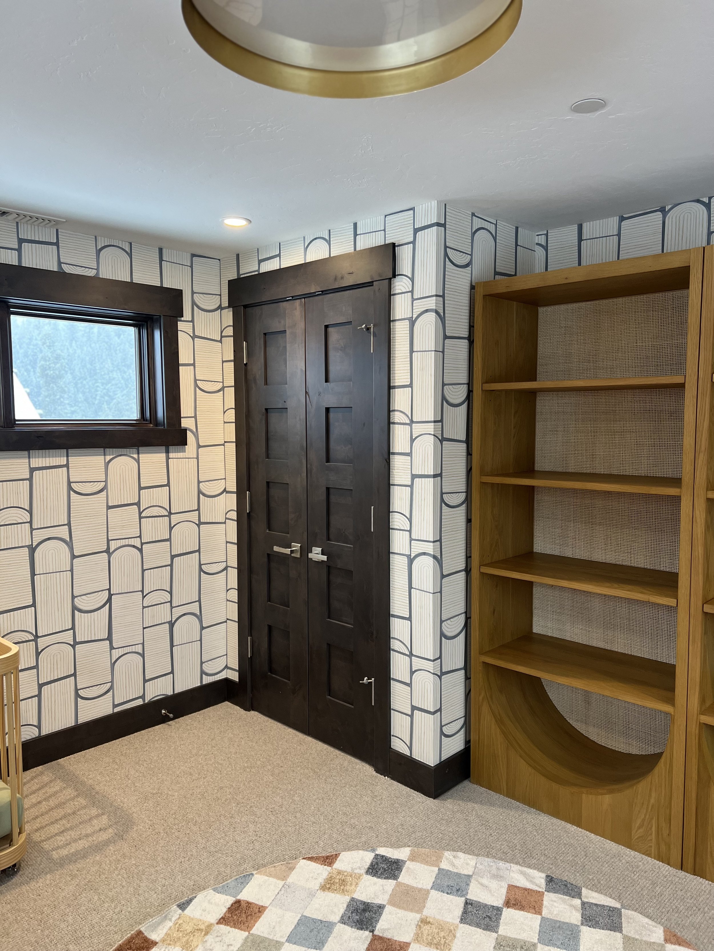 Room with modern geometric wallpaper, dark wood door, wooden shelving unit, crib, and a round patterned rug.