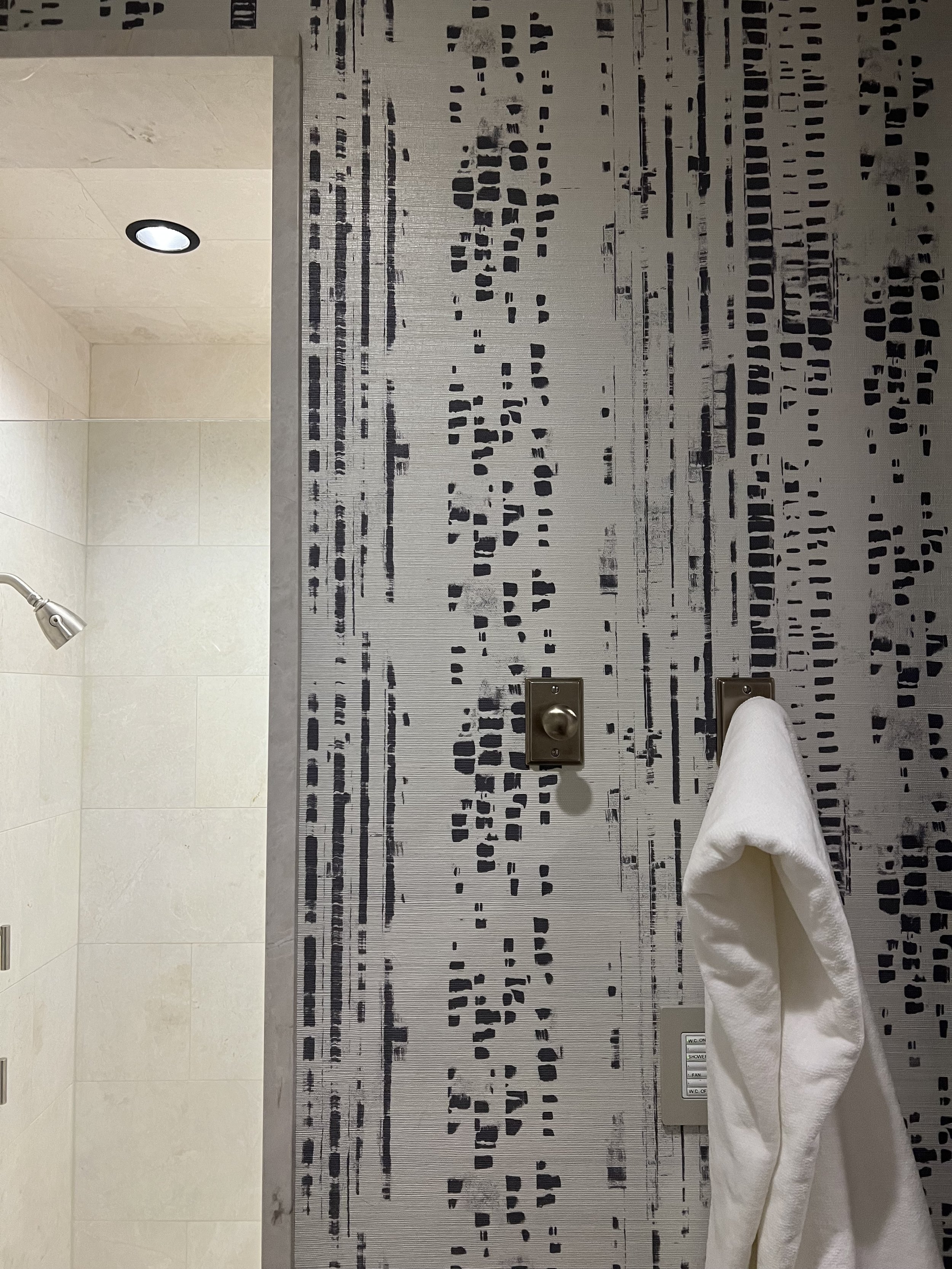 Bathroom wall with abstract patterned wallpaper, a mounted coat hook holding a white towel, and a glimpse of a tiled shower area with a showerhead and ceiling light.
