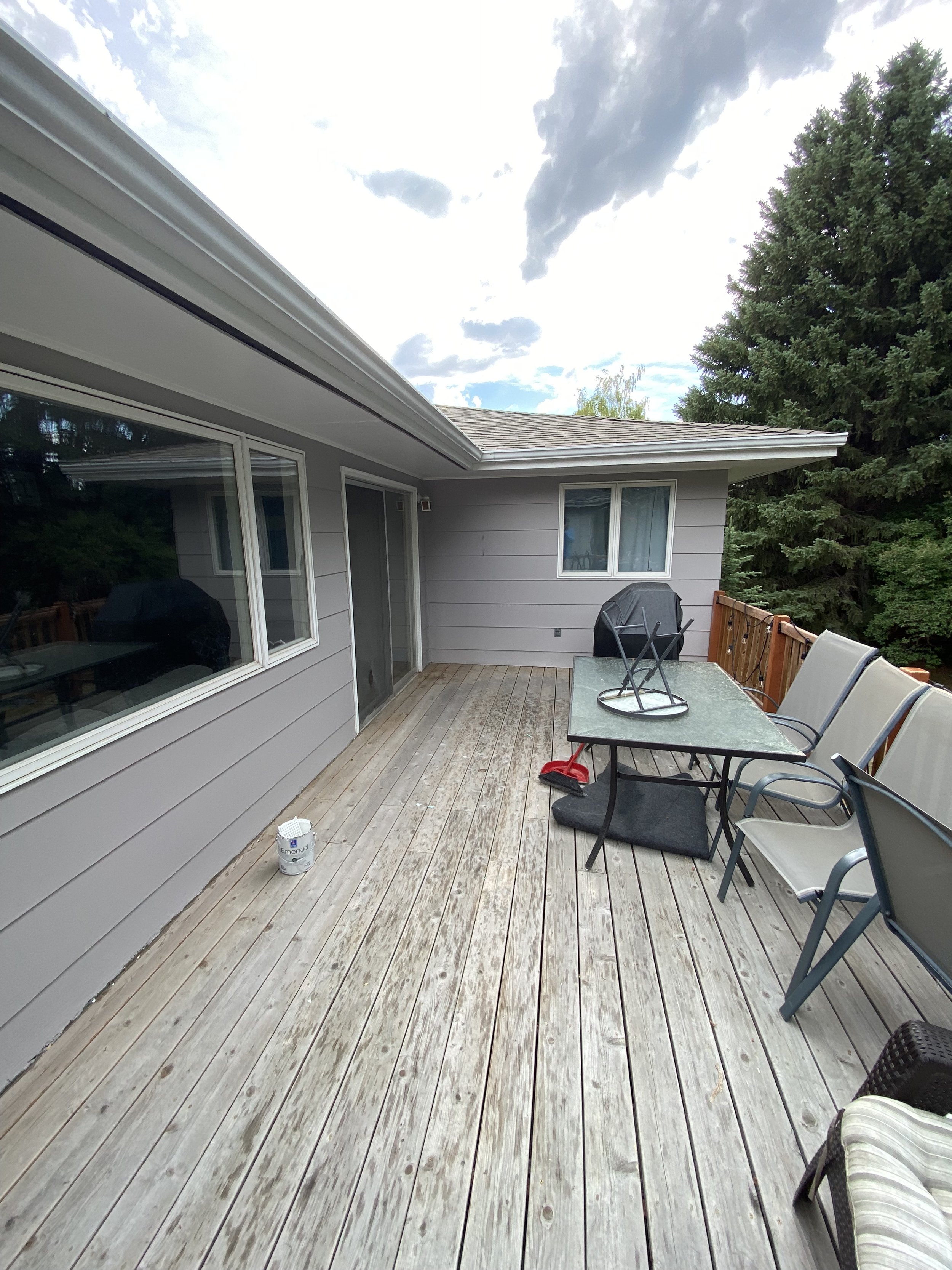 Wooden deck with outdoor furniture and a gray house exterior in the background. The deck has a table, chairs, an upside-down umbrella, and a paint bucket. Large tree and cloudy sky are visible.