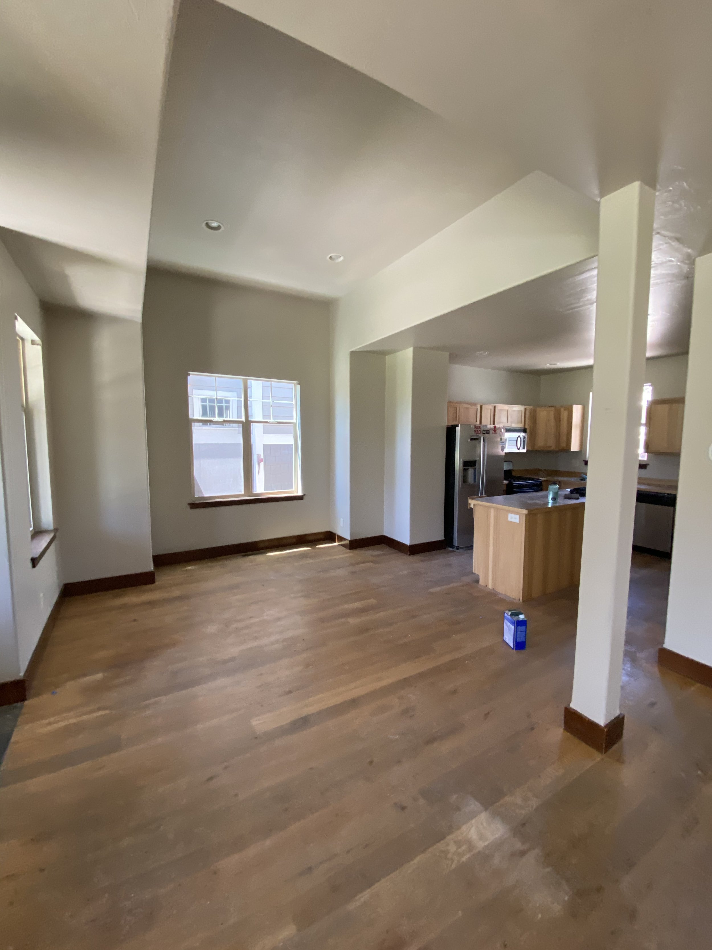 Empty living room with wooden flooring, adjacent to an open kitchen with light wood cabinets and stainless steel appliances.