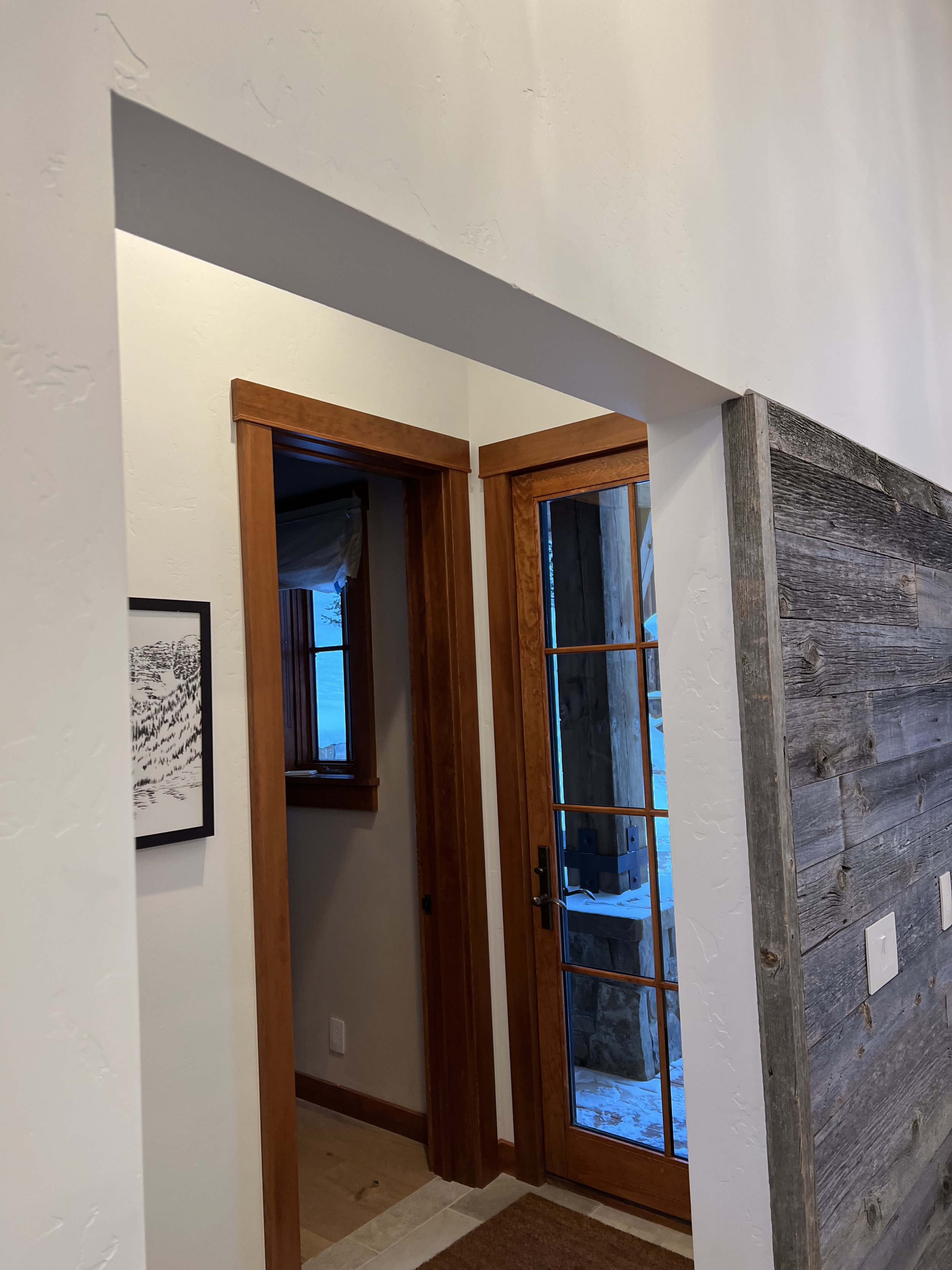 Interior of a hallway with wooden door frames and a glass door, partial view of a framed picture on the wall, and rustic wood paneling on one side. Bright natural light coming from the glass door.