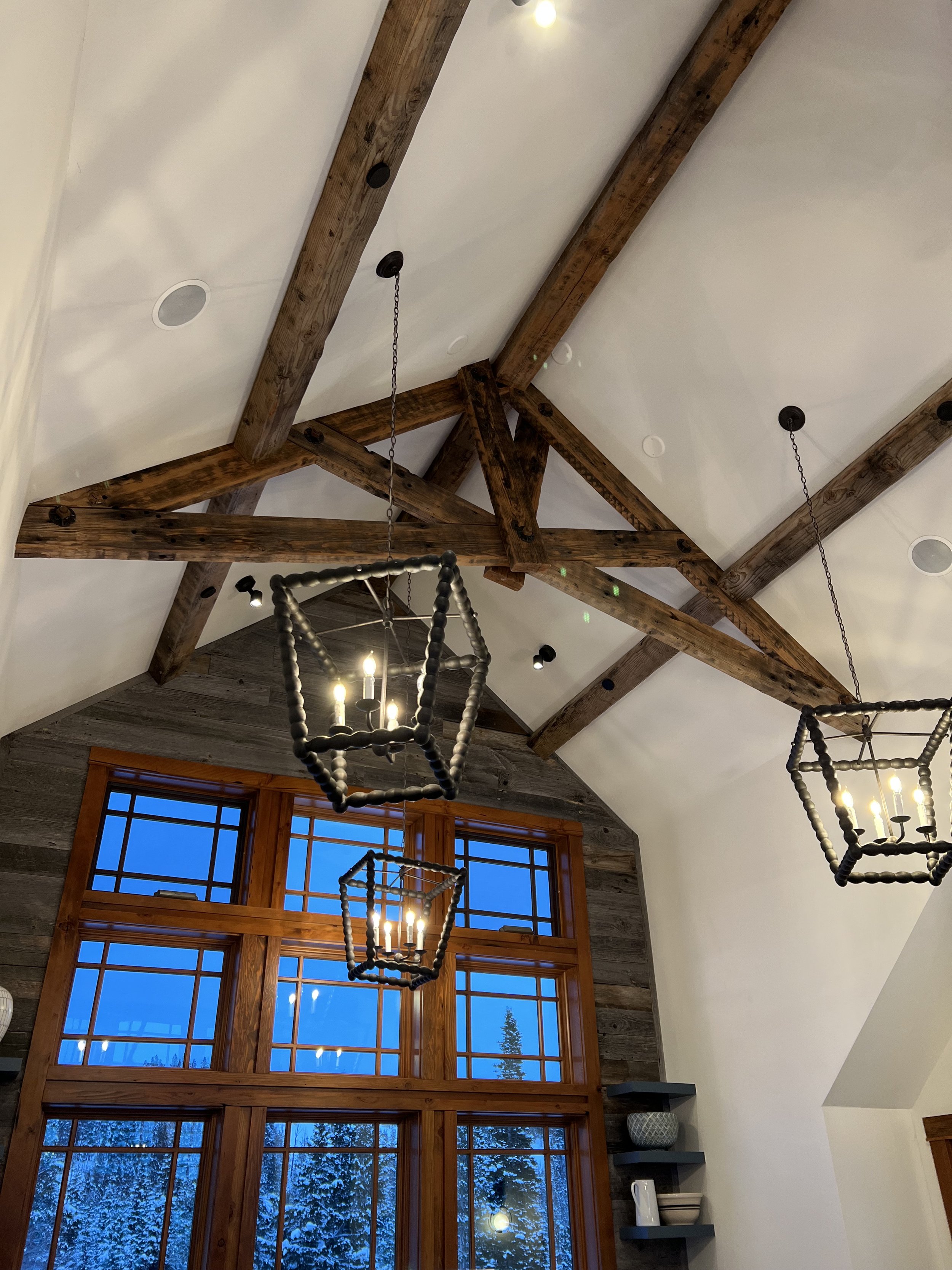 Interior ceiling view with exposed wooden beams and hanging black pendant lights. Large wooden-framed windows showcase a snowy outdoor scene with trees. Shelves with decorative items on the wall.