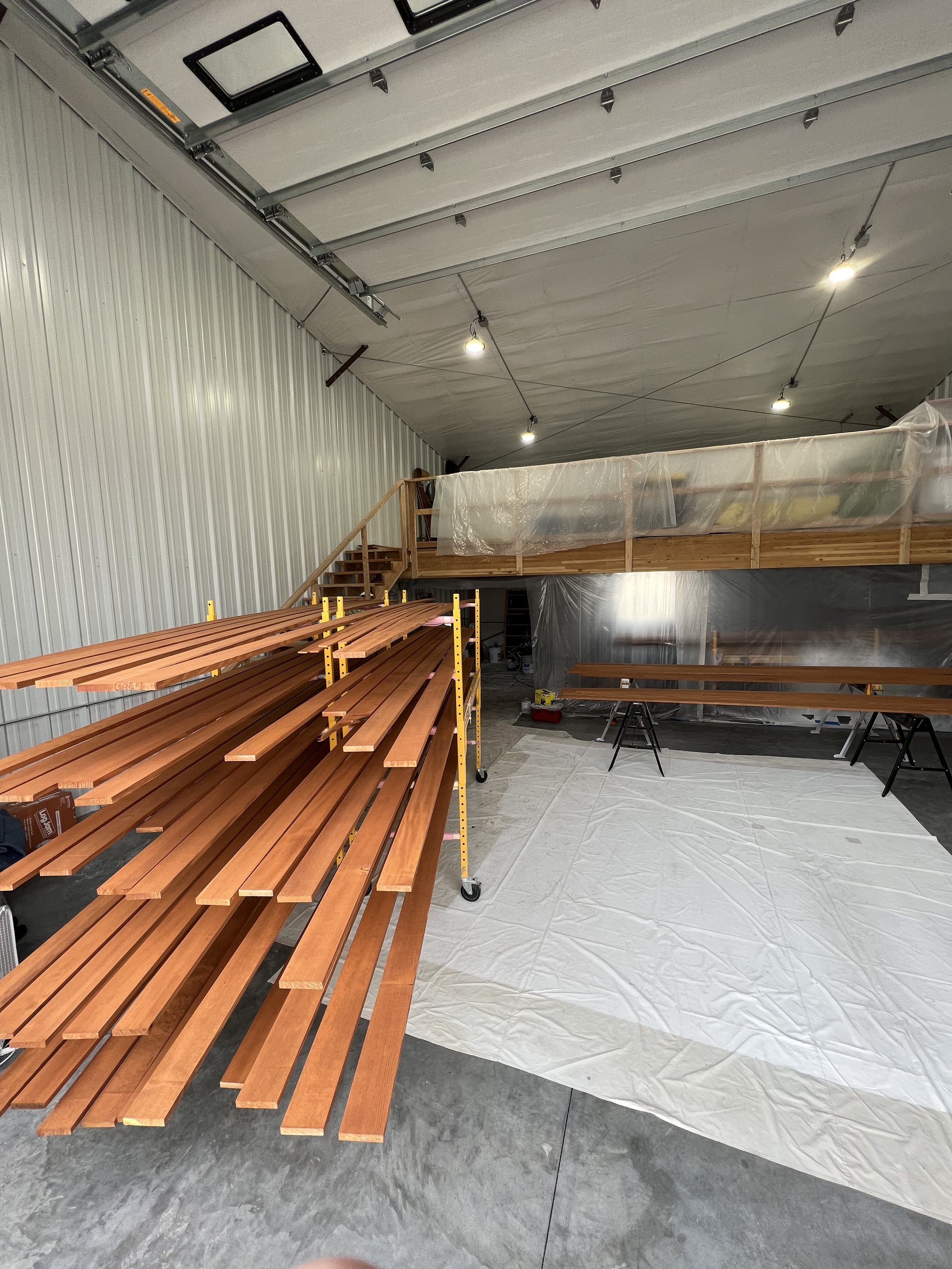 Interior of a workshop with stacked wooden planks, drop cloth, and an elevated work area with plastic covering.