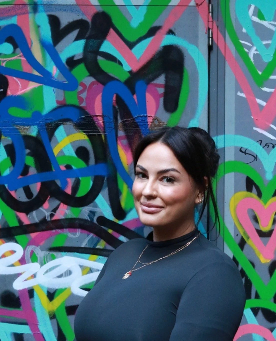 A woman with dark hair and a smile standing in front of a colorful graffiti wall with hearts and abstract shapes.