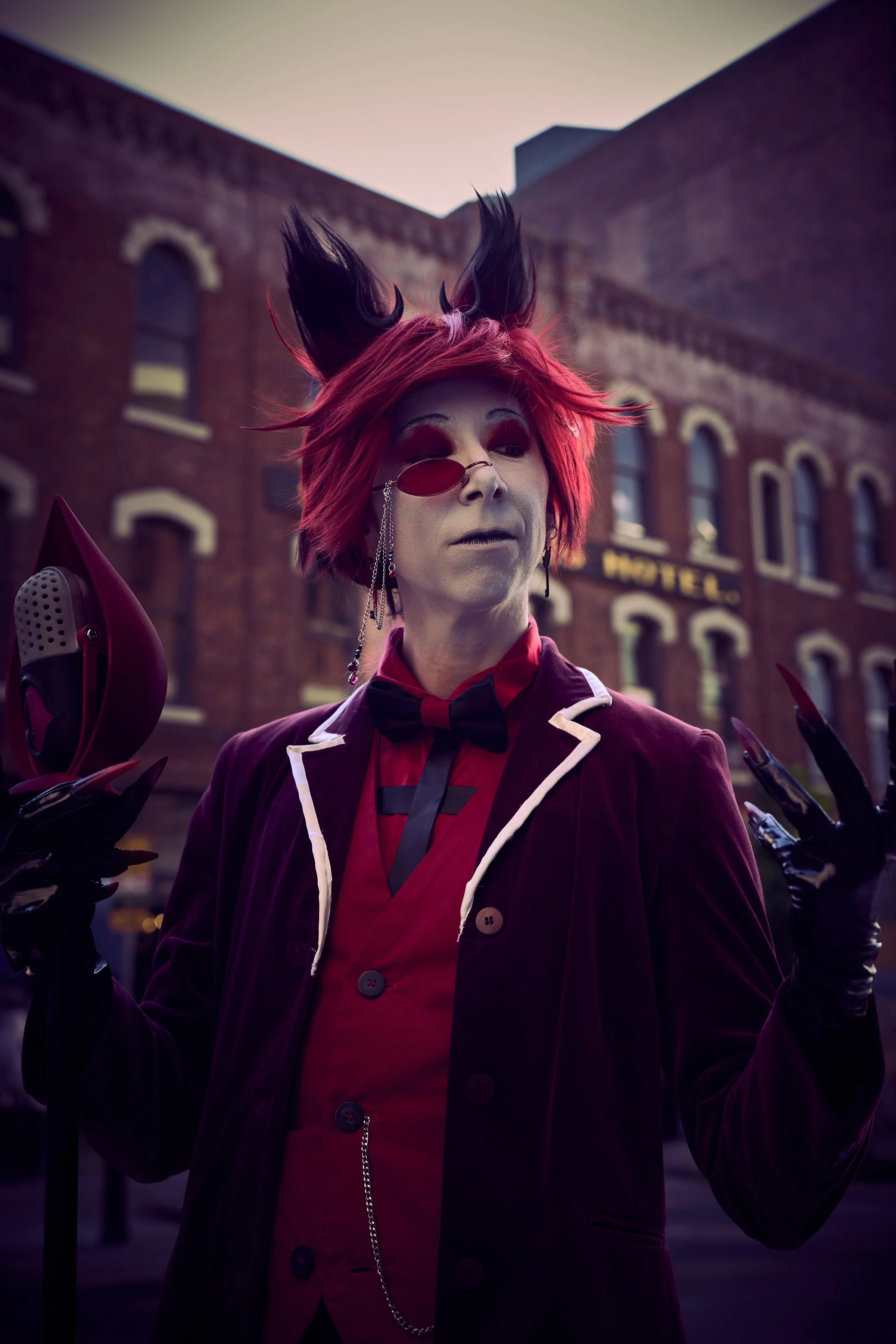 Person dressed in a red and black costume with a burgundy velvet jacket, red shirt, and black bow tie, wearing a red wig with black cat ears, holding a staff with a hat and wearing black gloves, in front of a brick building with a hotel sign at dusk.