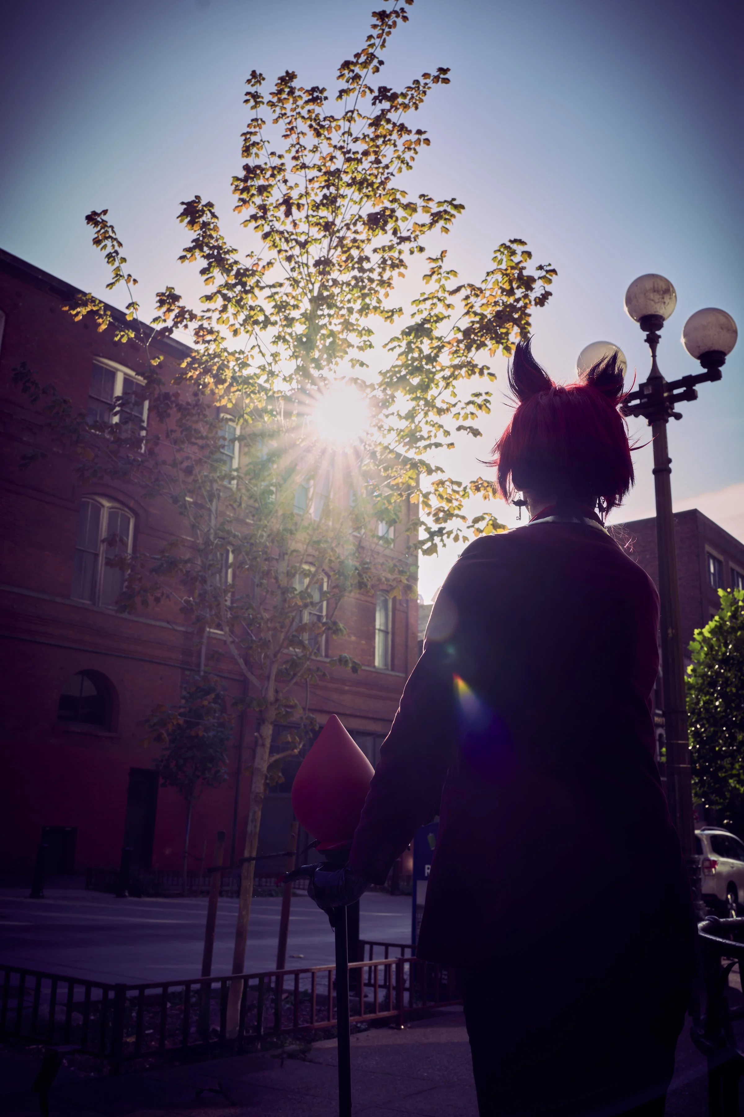 Person with red hair and fox ears holding a balloon, silhouetted against the sun in an urban street scene with buildings, trees, and streetlamps.