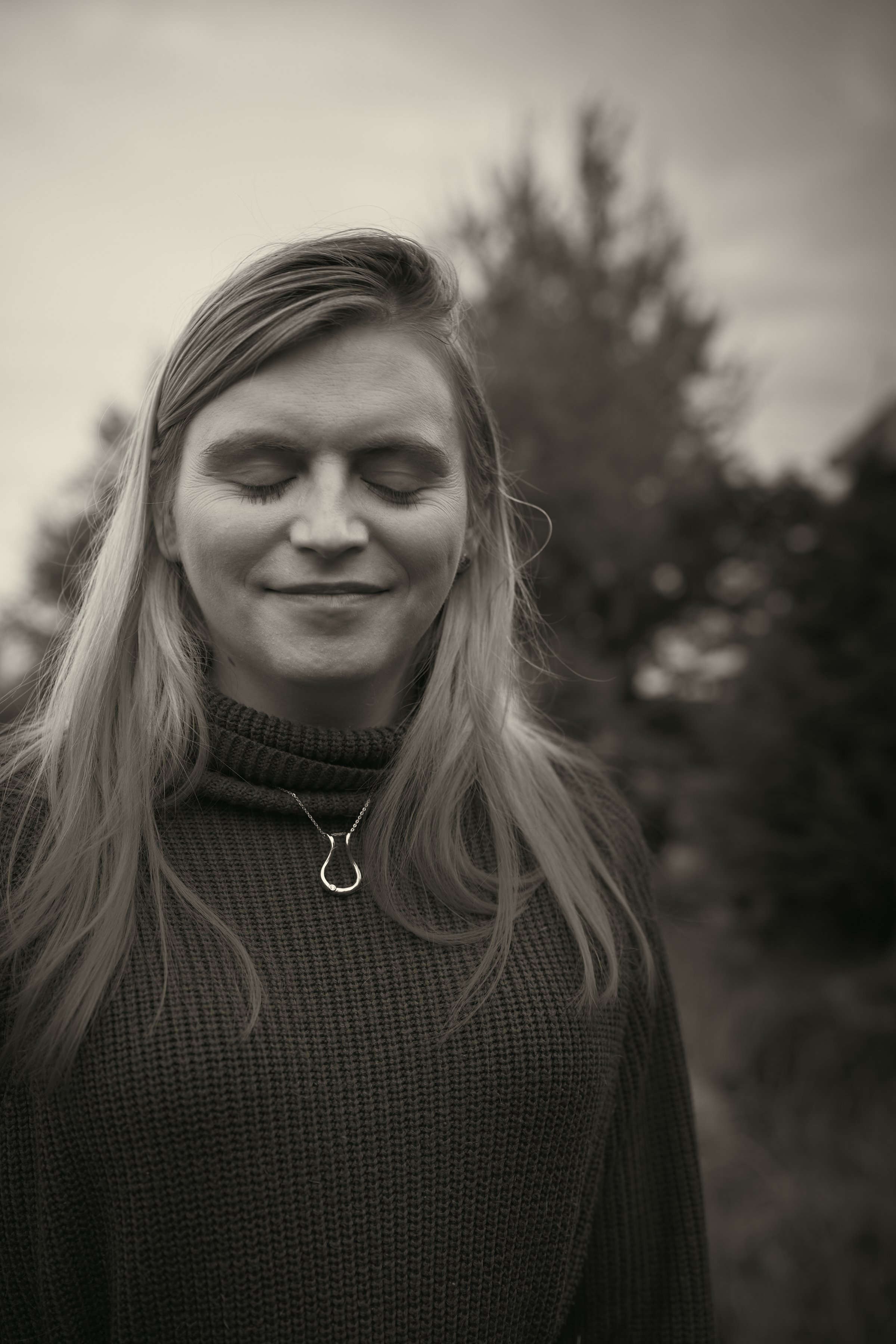 Black and white photograph of a woman with long blonde hair, smiling with her eyes closed, standing outdoors with a blurred background of trees.
