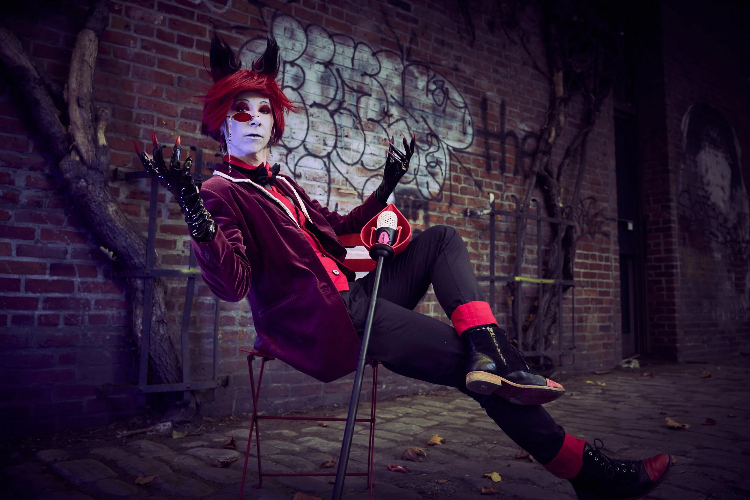 Person dressed in a dark red velvet blazer, black pants with red cuffs, black boots with red toe caps, and black gloves, sitting on a red metal chair in front of a graffiti wall with tree roots, holding a microphone, with red sunglasses and bright re