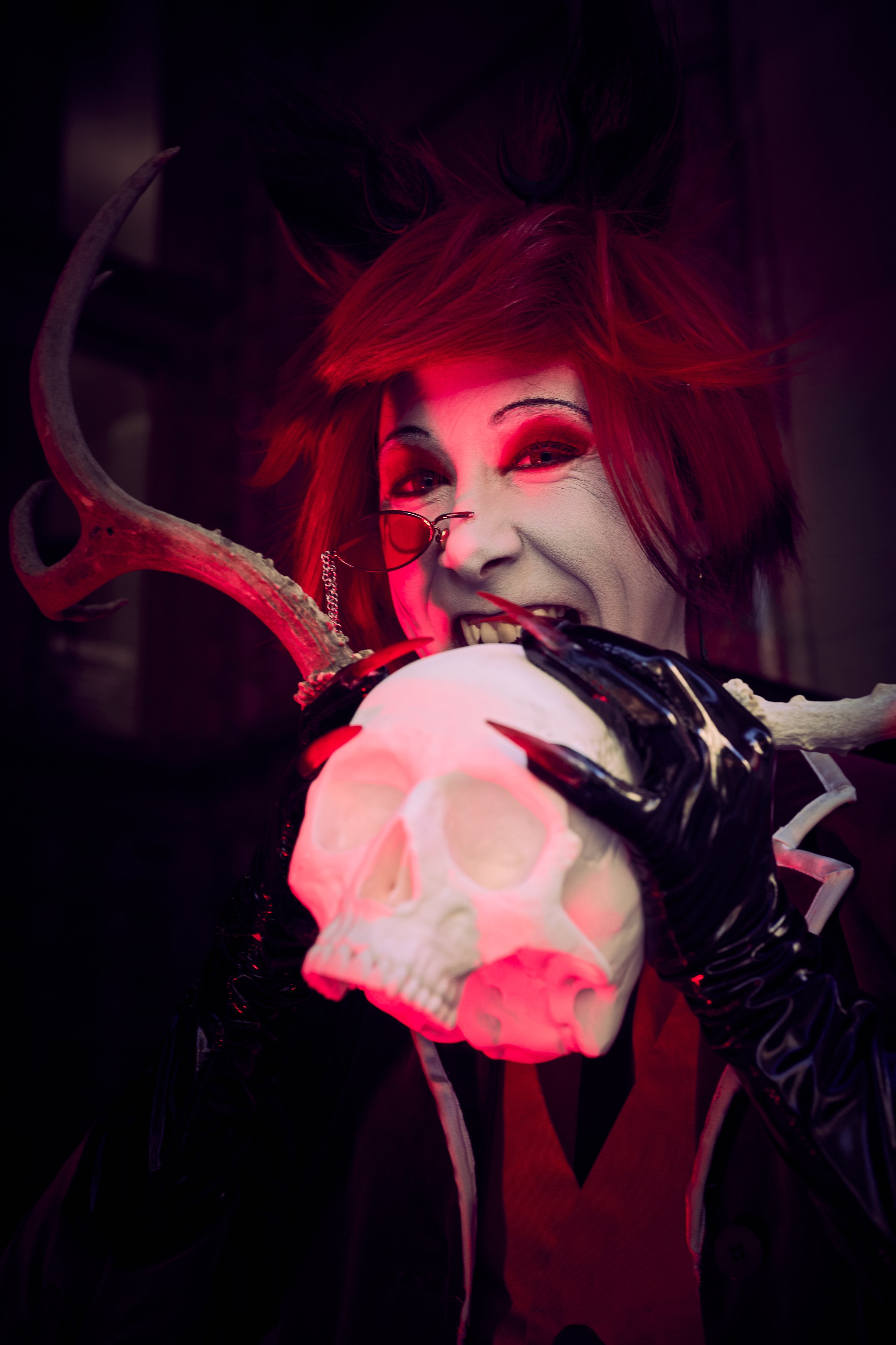A person dressed as a mischievous or evil character with red hair, wearing glasses, holding a white skull with horns, and smiling wickedly in a dark setting with red lighting.
