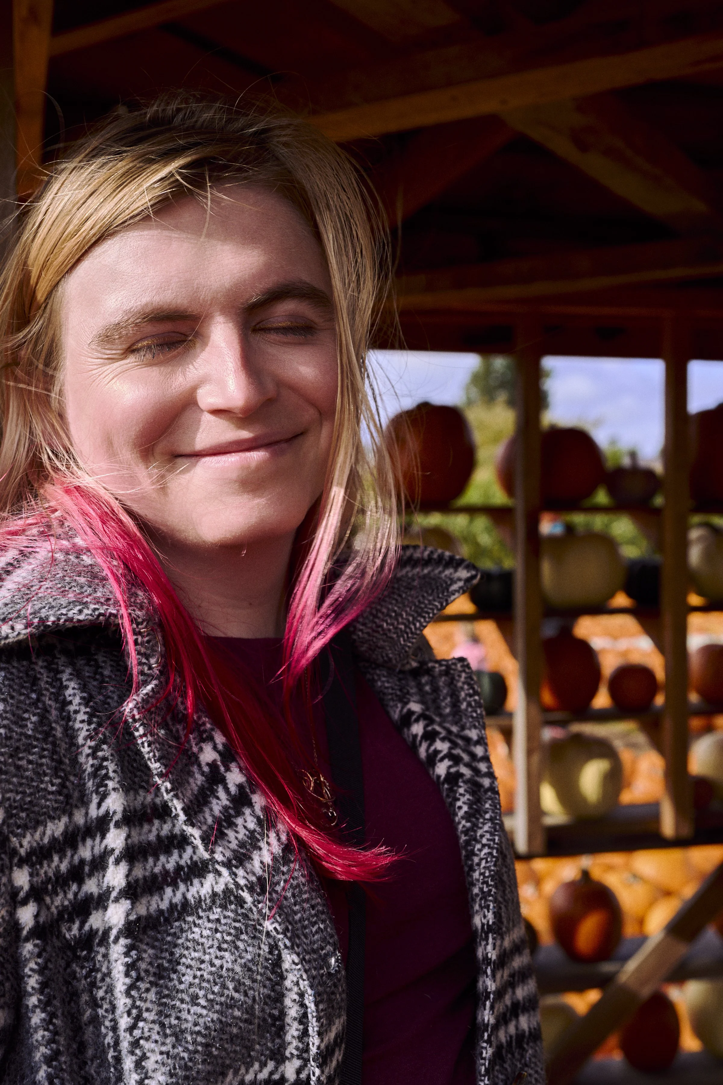 A woman with blond hair and pink streaks, wearing a black and white checkered jacket, smiling with closed eyes outdoors near a farm stand with pumpkins.