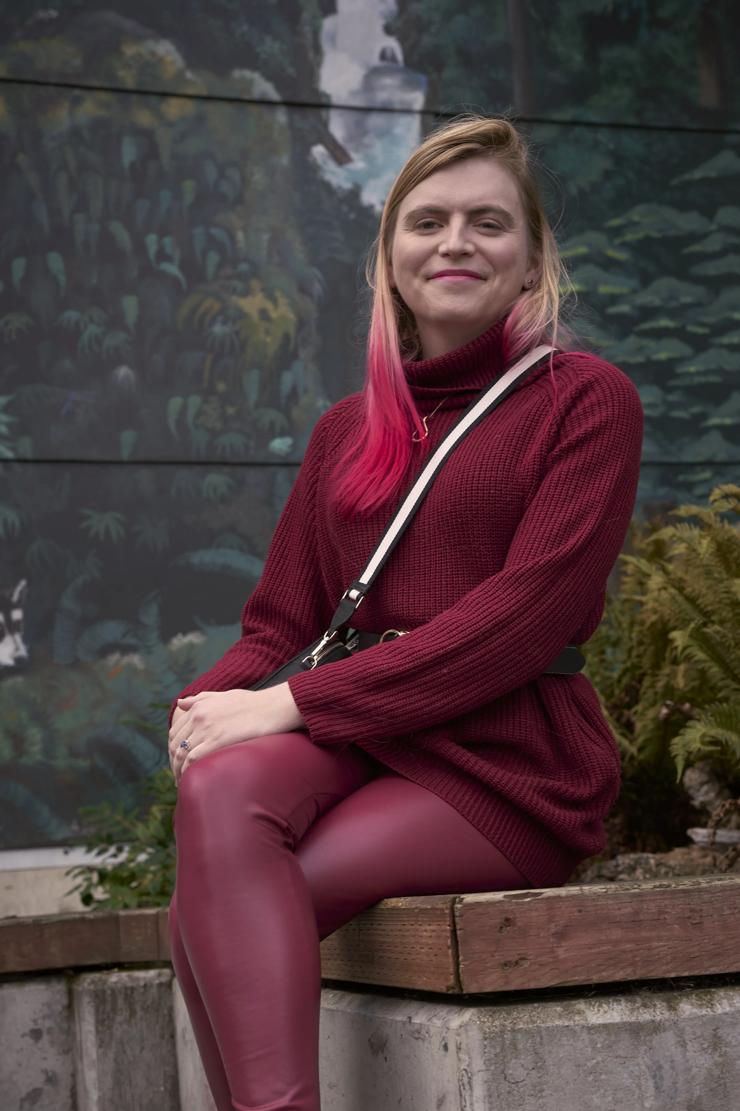 A woman with pink ombre hair, sitting on a wooden bench outdoors, wearing a maroon sweater and matching maroon pants, smiling at the camera with a colorful painted mural of greenery and water in the background.