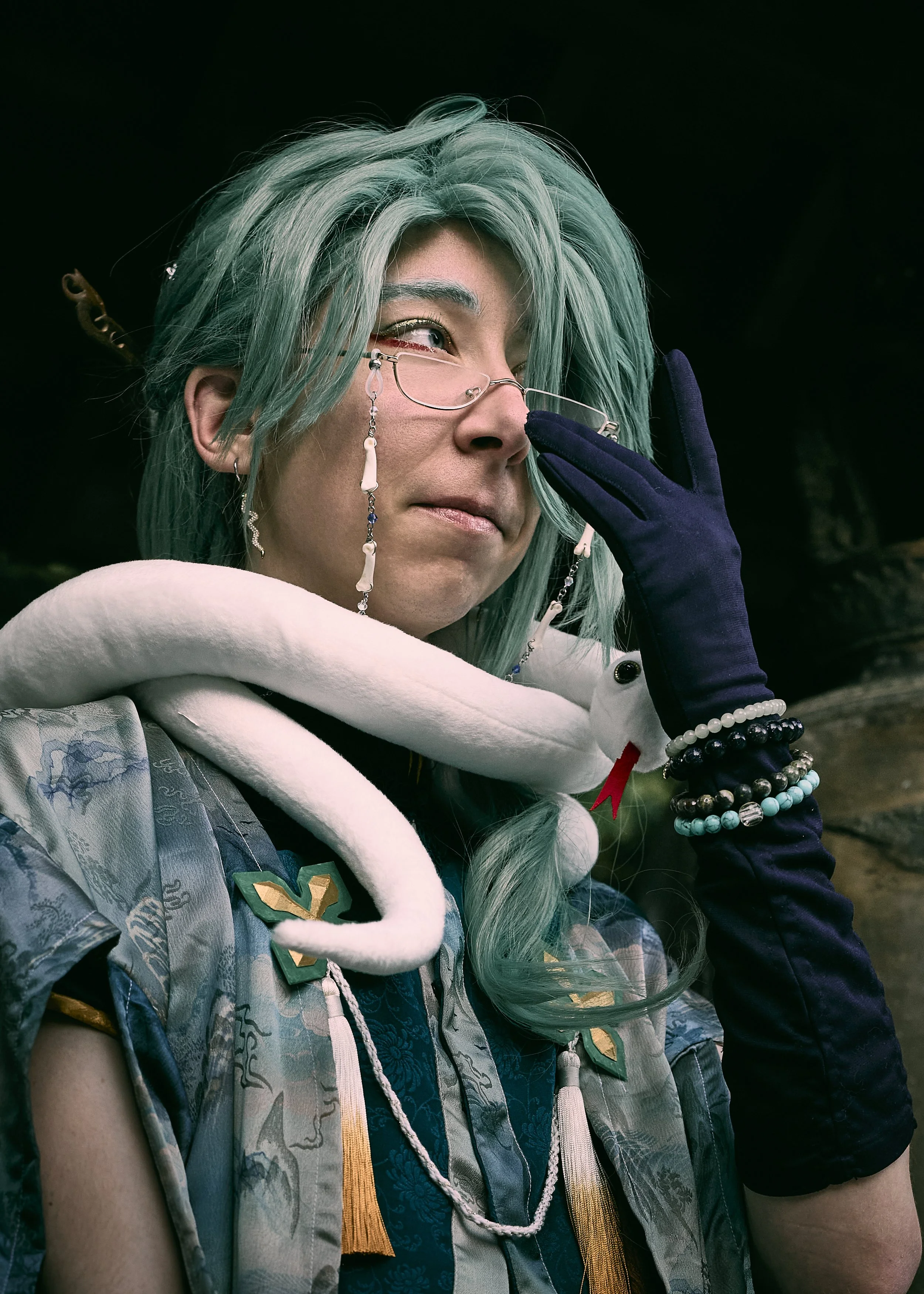 Person with long teal hair, glasses hanging from their face, multiple bracelets, wearing a snake-shaped scarf, outdoors with a dark background.