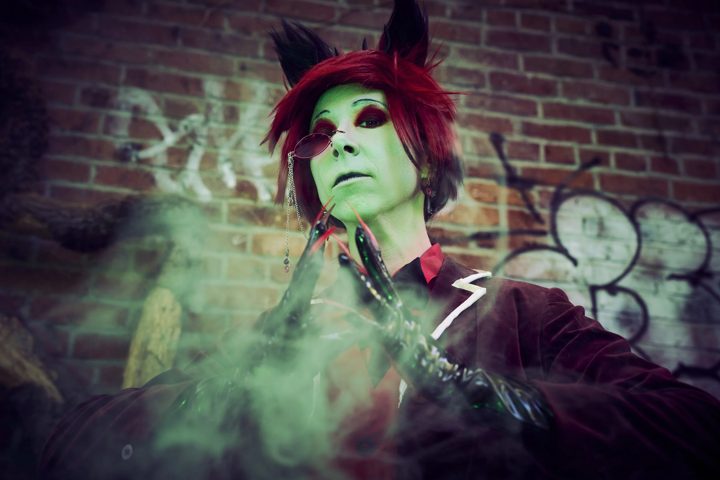 Person dressed as a cat demon with red hair, black cat ears, and green face paint, wearing a vampire costume with a black and white bow tie, posing with black claws in front of a graffiti-covered brick wall.
