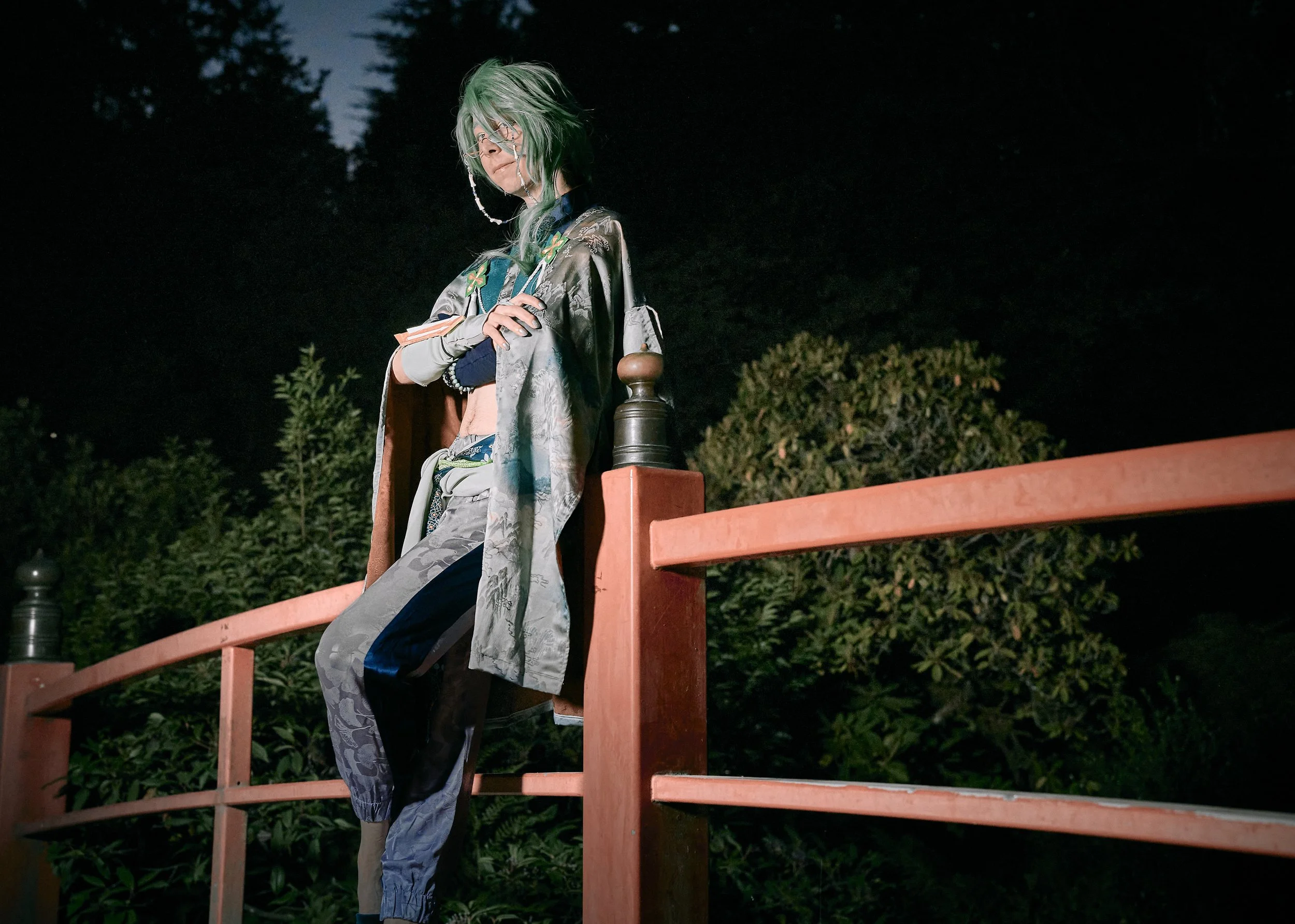 A person with green hair, glasses, and tattoos standing on a red bridge at night. They are wearing layered clothing, including a kimono-style jacket, a crop top, and baggy pants, with a serious expression.