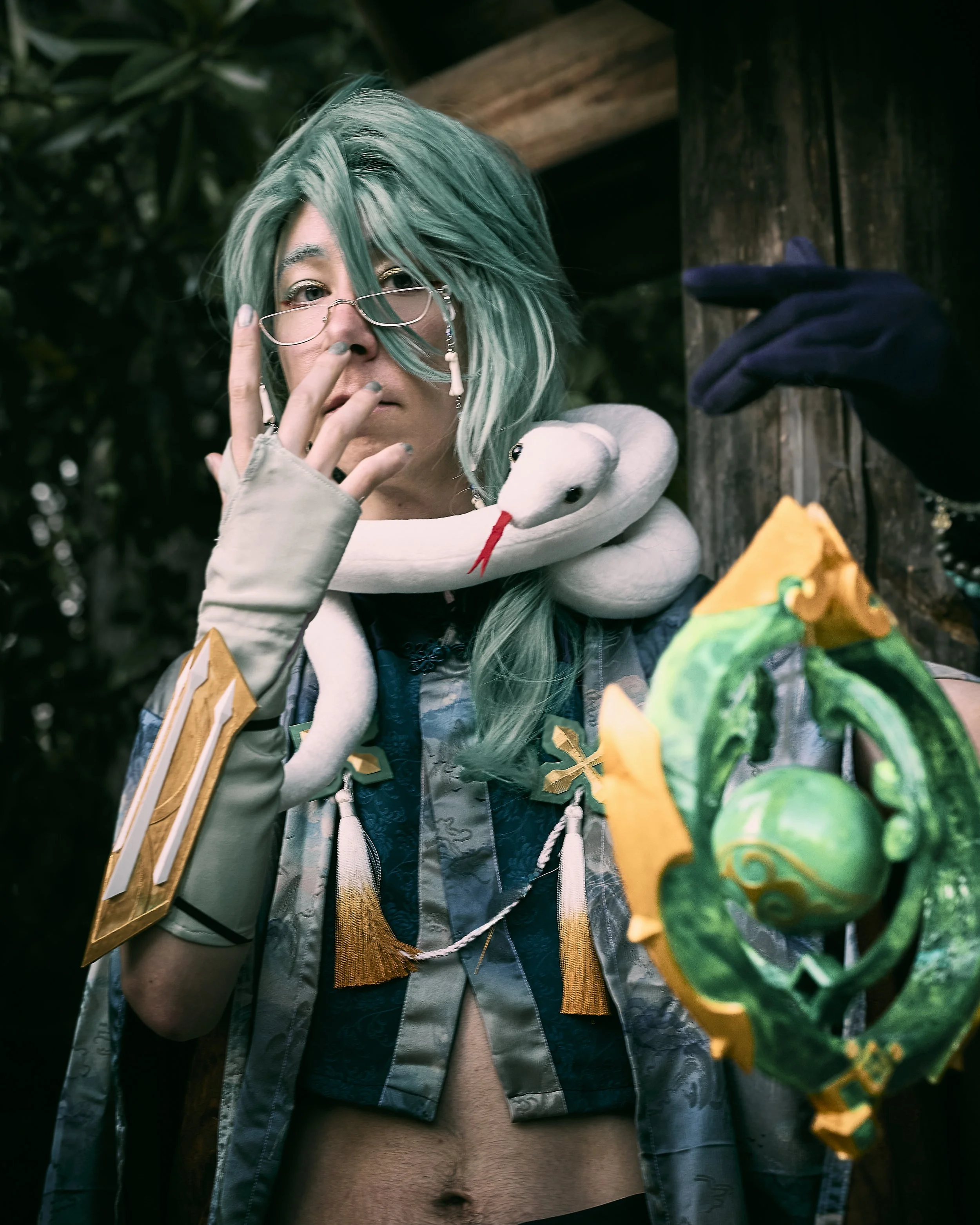 A person with teal hair and glasses wears a snake around their neck, a patterned cloak, and holds a colorful staff, standing outdoors near wooden structures and foliage.