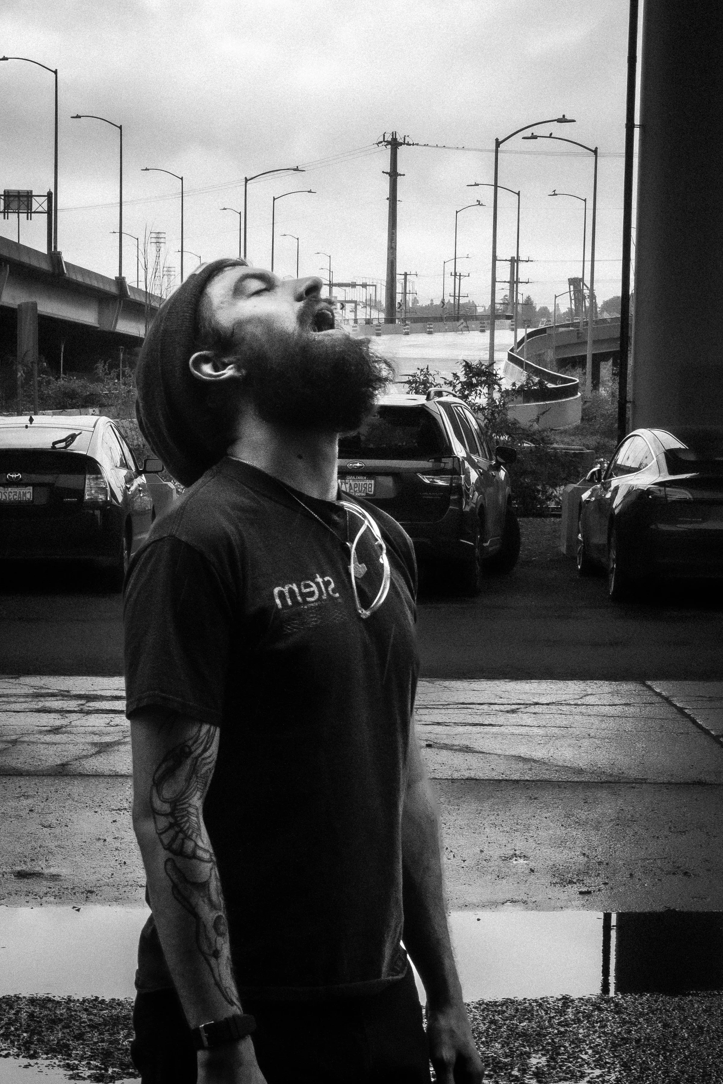A man with a beard and tattoos on his arm, wearing a dark T-shirt and a head covering, stands outdoors on a cloudy day with his head tilted back and eyes closed, possibly yawning or singing, with cars and power lines in the background.