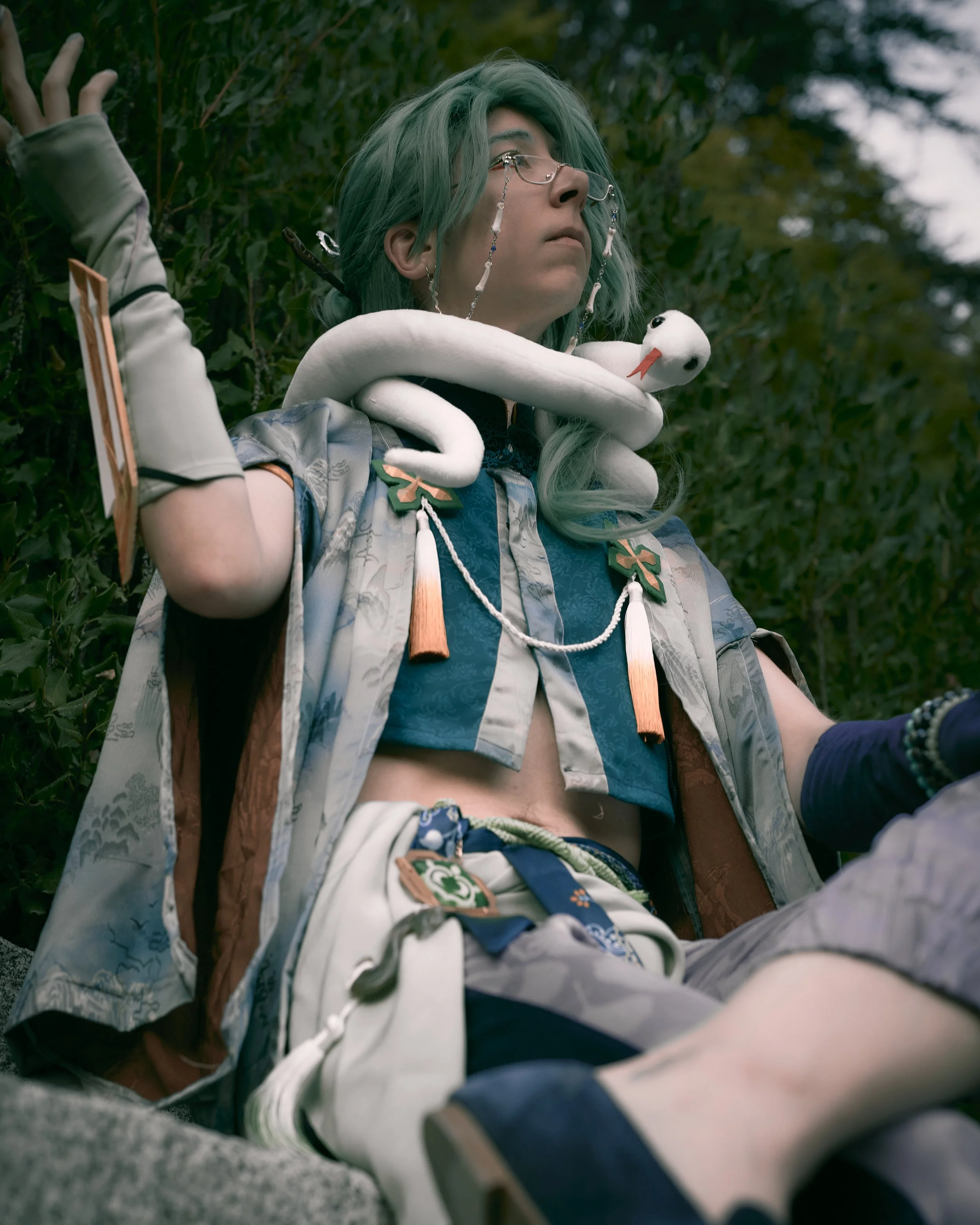 Person with green hair, glasses, and ornate clothing sits outdoors with a white toy swan around their neck, surrounded by greenery.