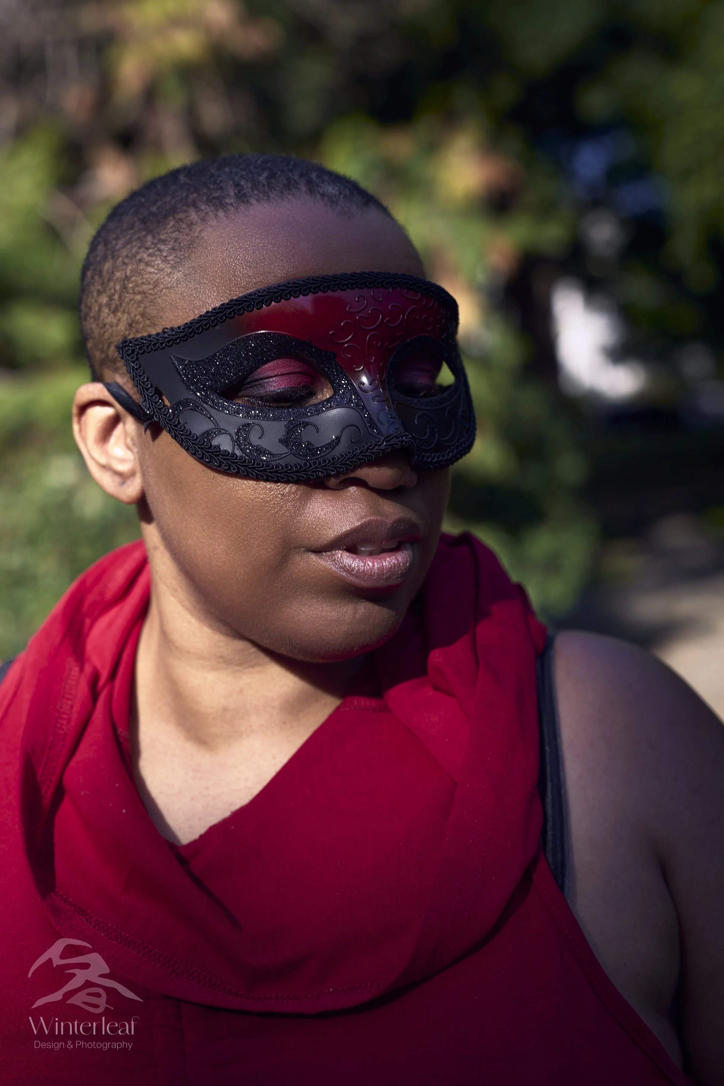 A woman with short hair wearing a decorative black and red mask and a red sleeveless top outdoors with green trees in the background.