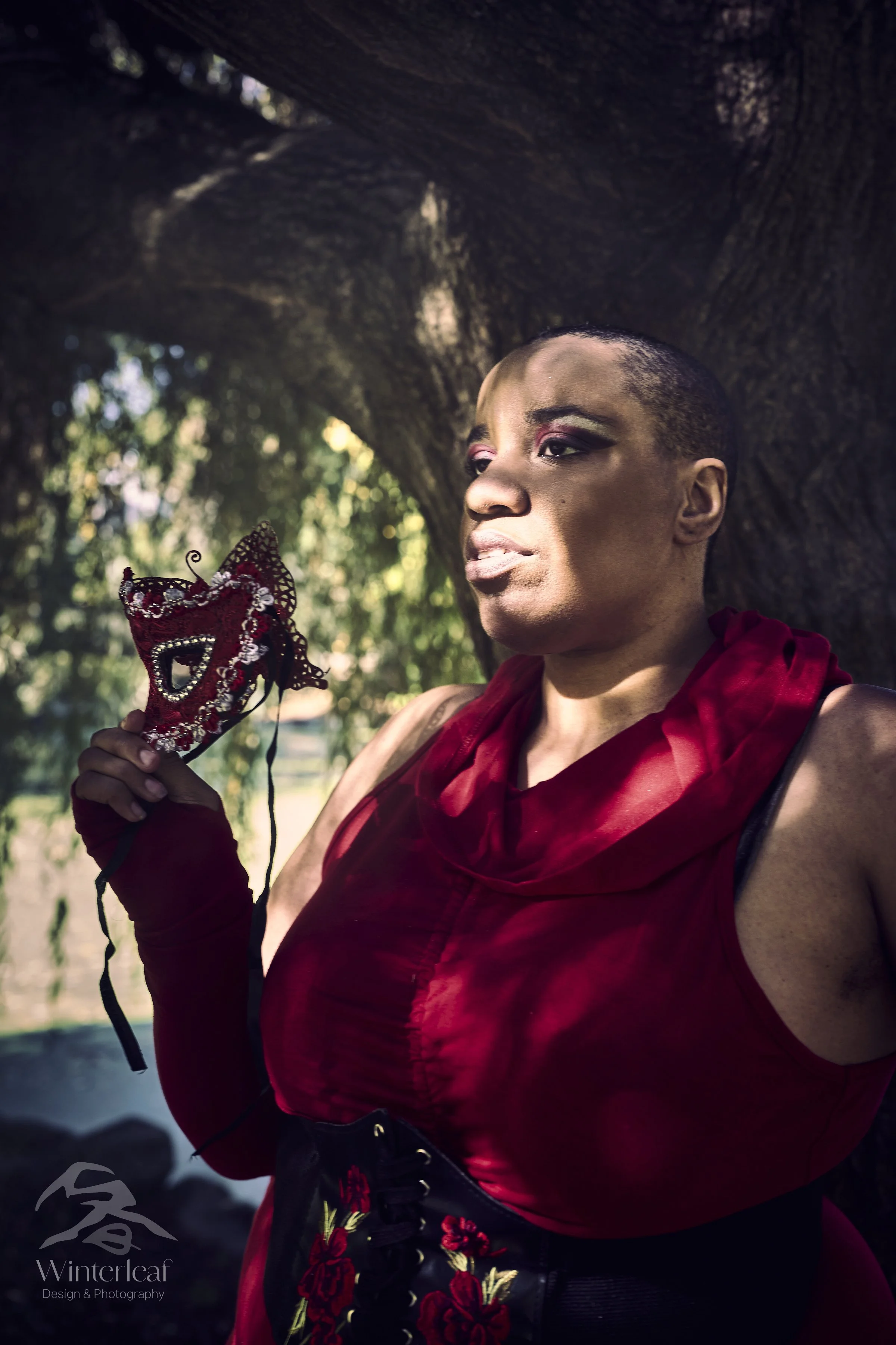Person holding a red and black decorative mask outside near a large tree, with sunlight filtering through leaves.