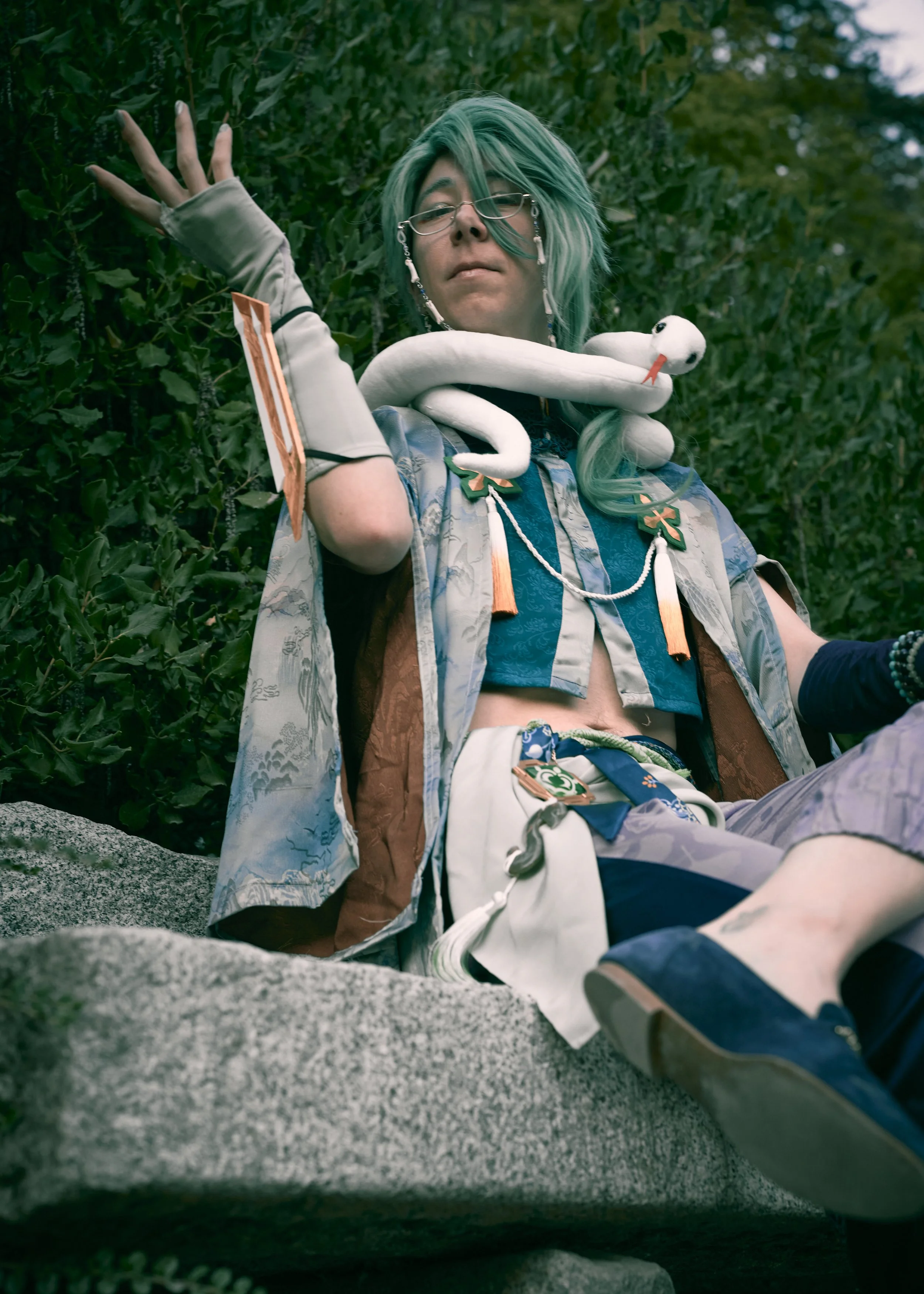 A person with teal hair, glasses, and outdoor clothing sitting outdoors with a white plush swan perched on their shoulder, holding colorful ornaments, with greenery in the background.