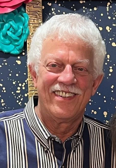 A smiling older man with white hair and a mustache, wearing a striped shirt, standing in front of a decorated wall with colorful, textured art pieces and gold speckles.
