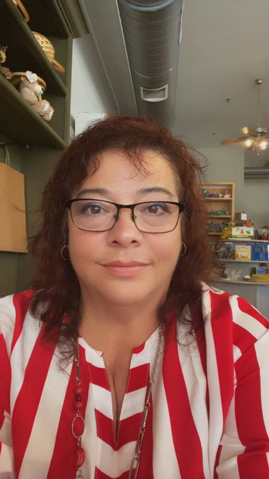 A woman with glasses and earrings, wearing a red and white striped top, taking a selfie in an indoor setting with shelves and decorations in the background.