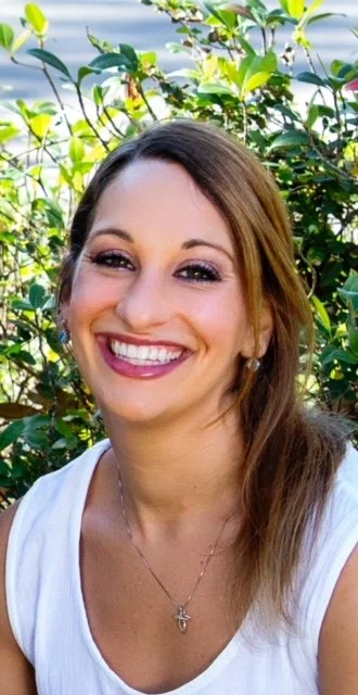 Smiling woman with long brown hair and earrings, wearing a white top and a cross necklace, outdoors with green foliage in the background.