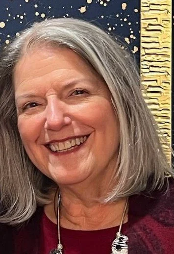 Close-up of a smiling woman with shoulder-length gray hair, wearing a burgundy top and a necklace, with a decorative black and gold background.