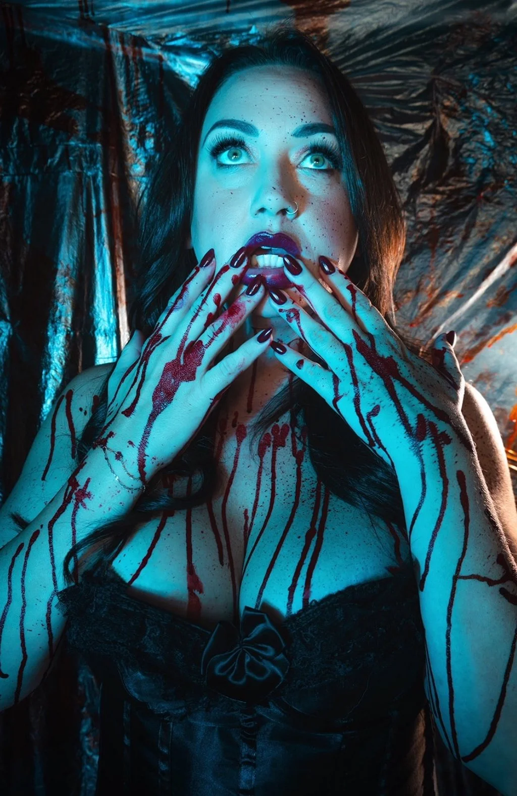 A woman with dark hair and bright eyes, covered in fake blood, with blood on her hands and face, and wearing a black dress with a bow, standing in front of a metallic background.