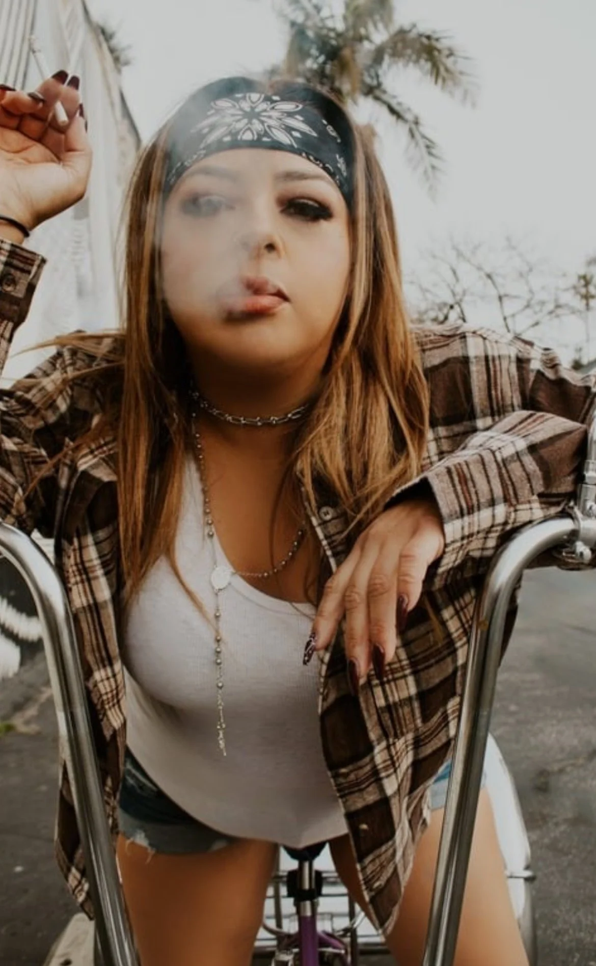 A young woman with long hair, wearing a plaid shirt, white top, black shorts, and jewelry, sitting on a motorcycle outdoors with a bandana on her head, looking through a motorcycle windshield.