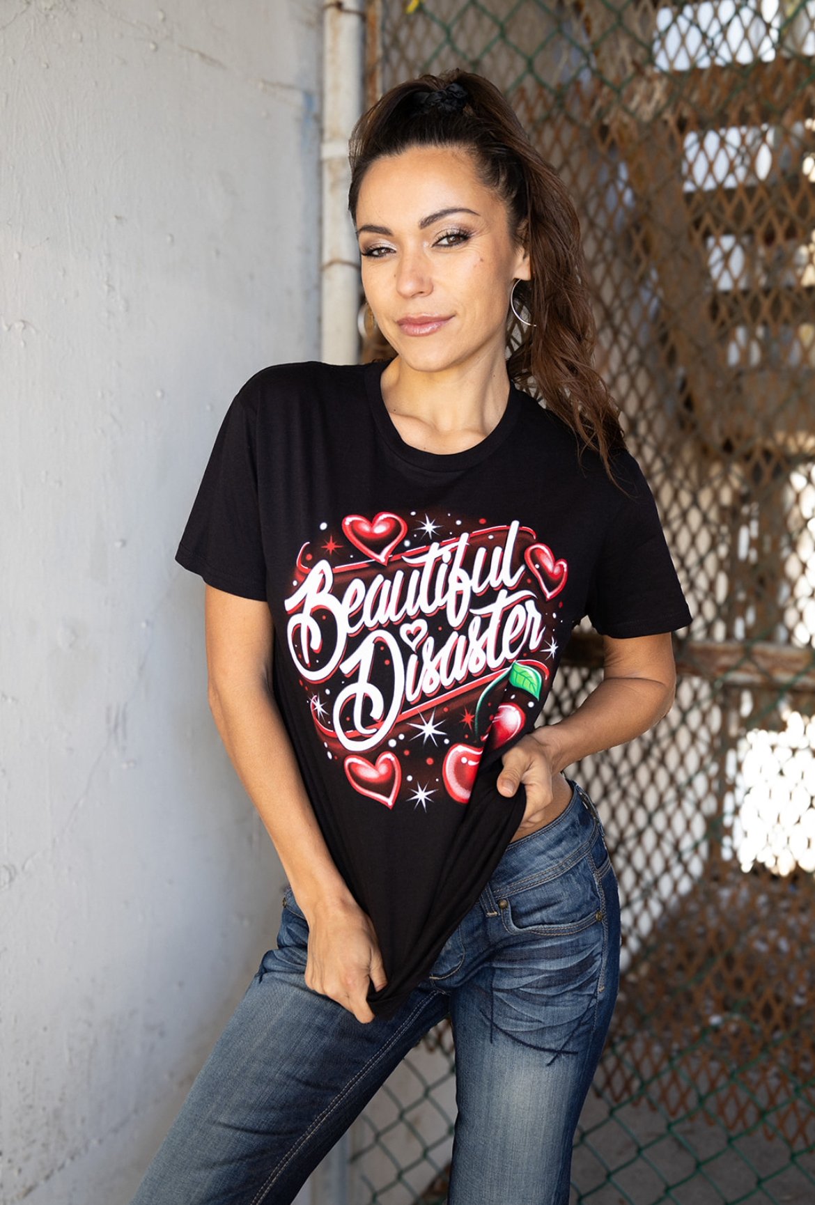 A woman wearing a black T-shirt with the words "Beautiful Disaster" and heart graphics, standing against a fence and wall.